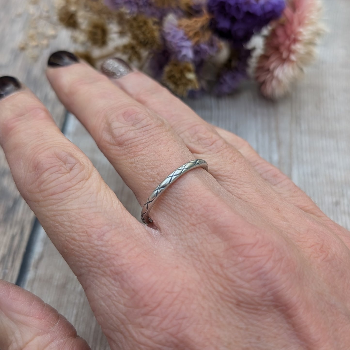 Delicate silver ring with a crosshatch pattern called "Starlight," shown on a person’s finger, with a subtle background of wood and dried flowers.