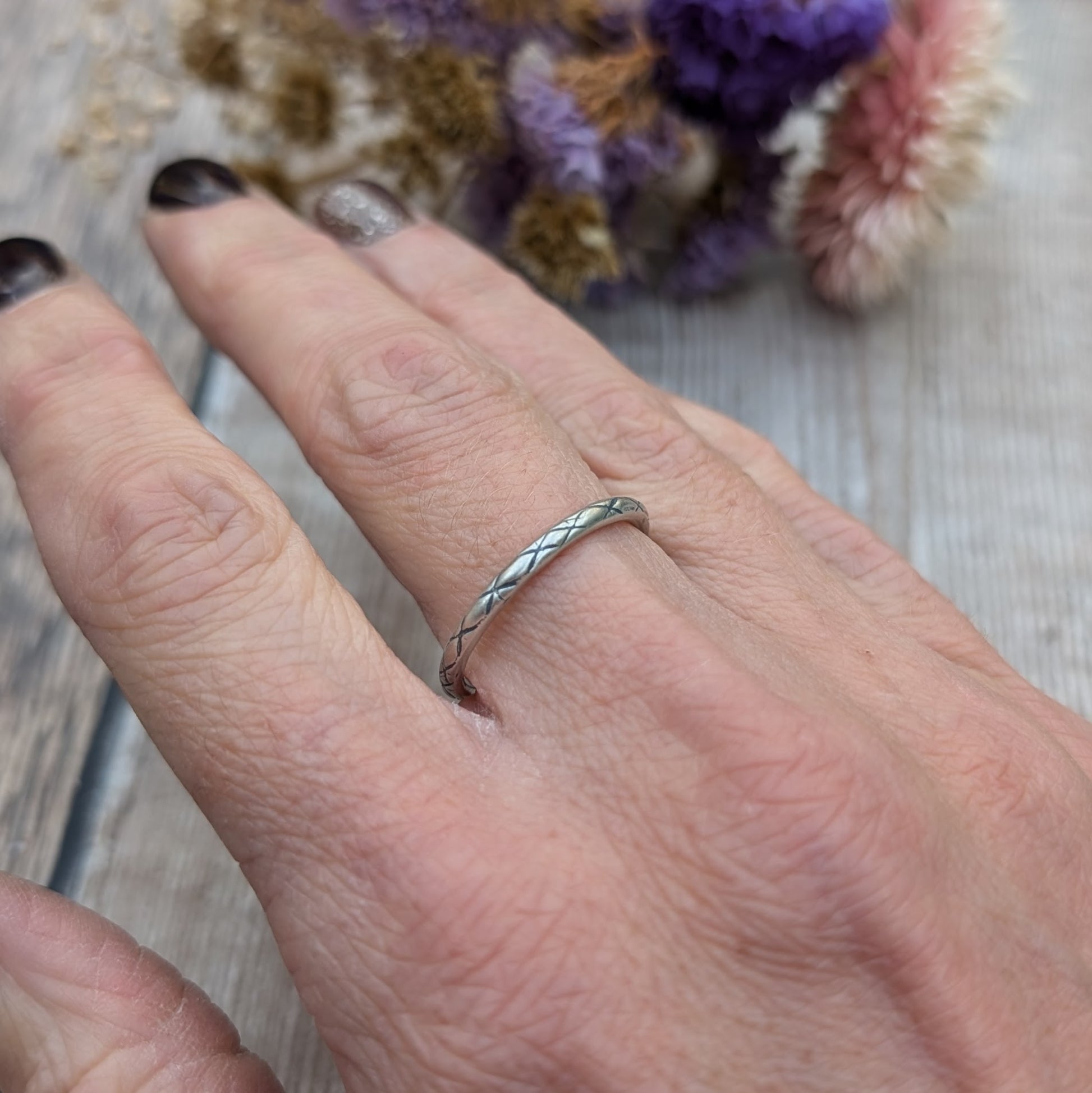 Delicate silver ring with a crosshatch pattern called "Starlight," shown on a person’s finger, with a subtle background of wood and dried flowers.