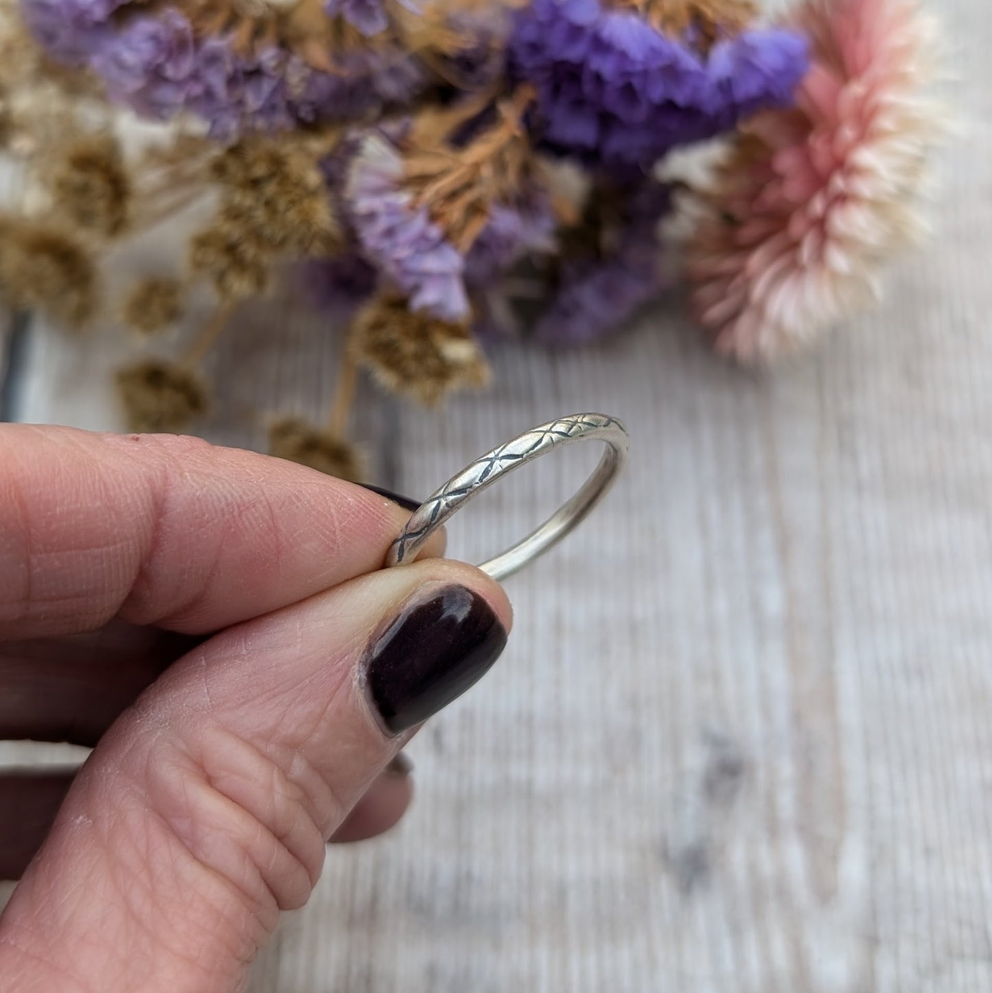 Close-up of a hand holding a delicate silver ring with a crosshatch pattern called "Starlight," set against a soft background of dried flowers in purple and pink hues.