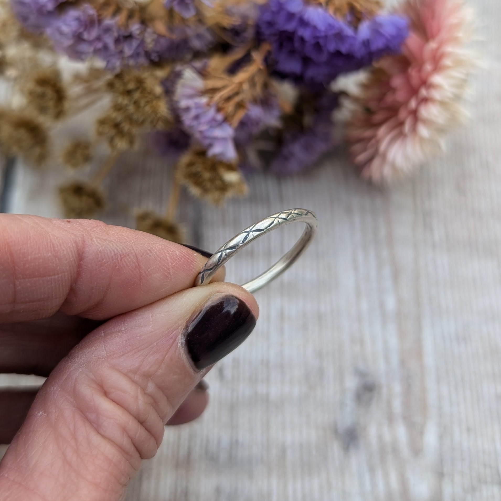 Close-up of a hand holding a delicate silver ring with a crosshatch pattern called "Starlight," set against a soft background of dried flowers in purple and pink hues.