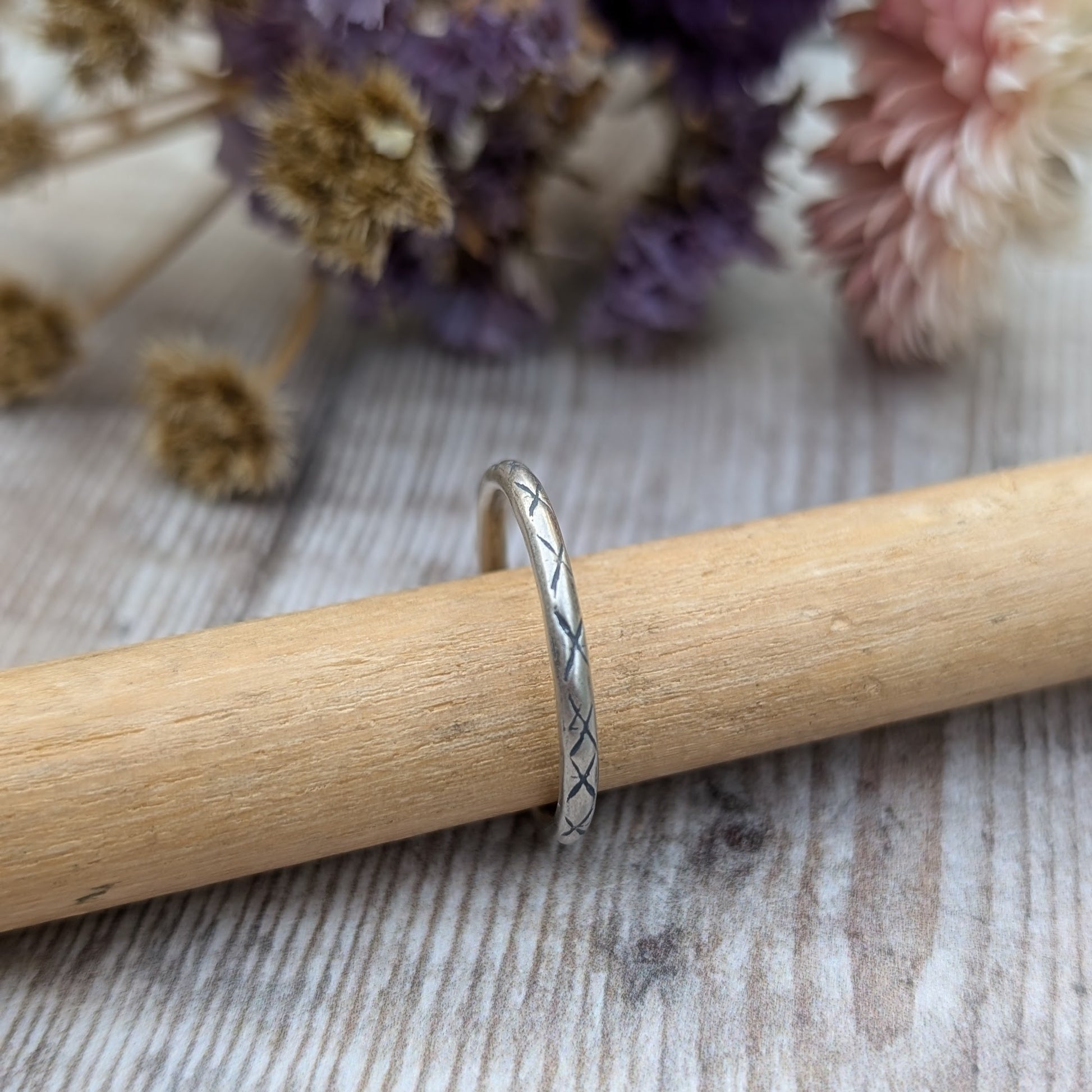 Silver ring with a crosshatch pattern called "Starlight," displayed on a wooden dowel, with dried flowers in soft purple and beige tones in the background.