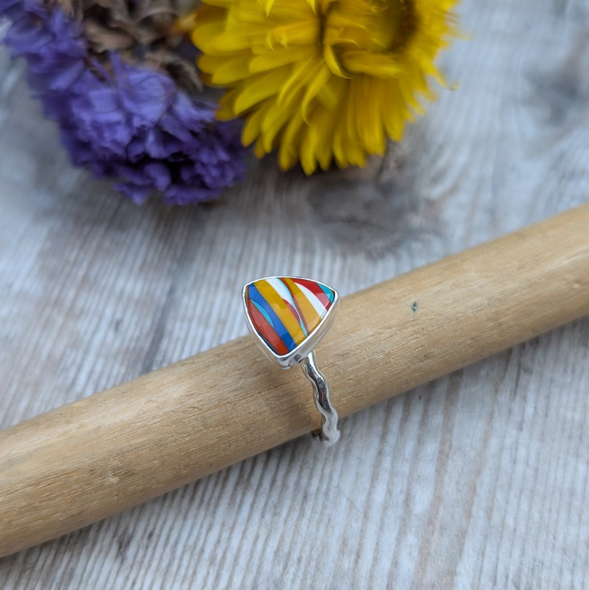 The Surfite triangle ring displayed on a wooden ring mandrel with bright yellow and purple flowers in the background.