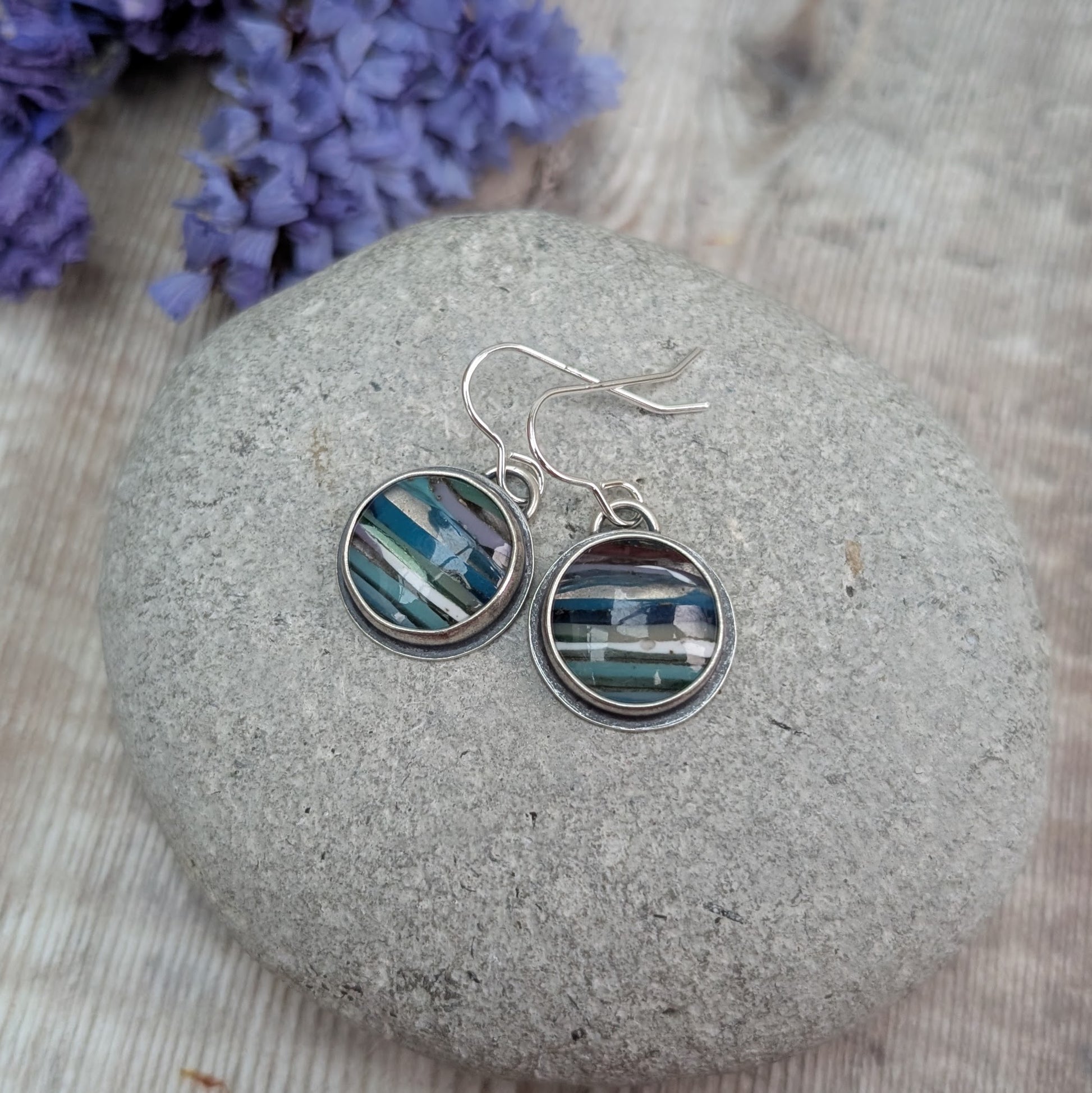Each earring is a round surfite stone set in an oxidised silver bezel, suspended from silver earring wire. Surfite has random horizontal uneven stripes of lilac, blue, turquoise, green, white and black, gloss finish.
