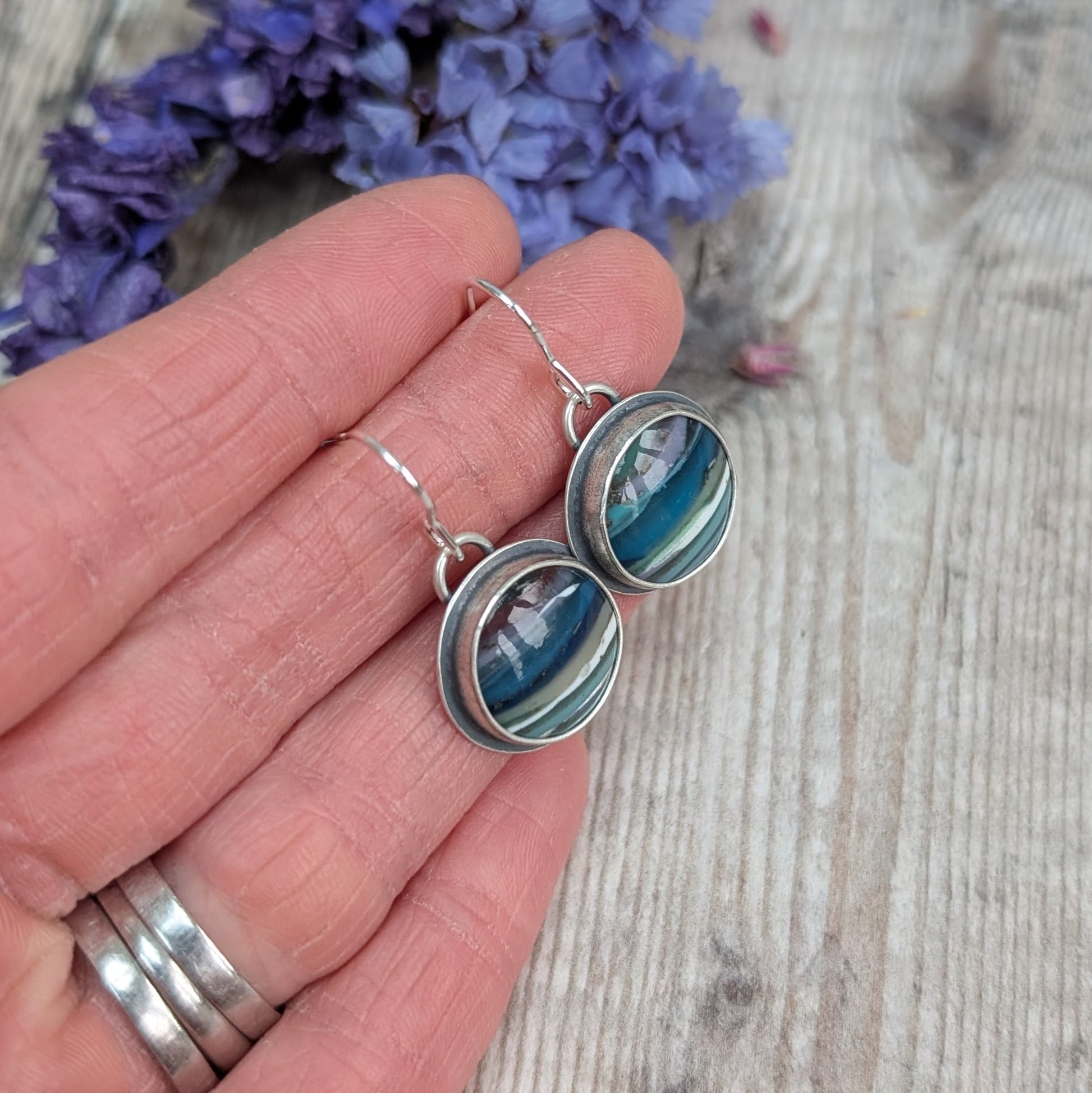 Each earring is a round surfite stone set in an oxidised silver bezel, suspended from silver earring wire. Surfite has random horizontal uneven stripes of lilac, blue, turquoise, green, white and black, gloss finish.