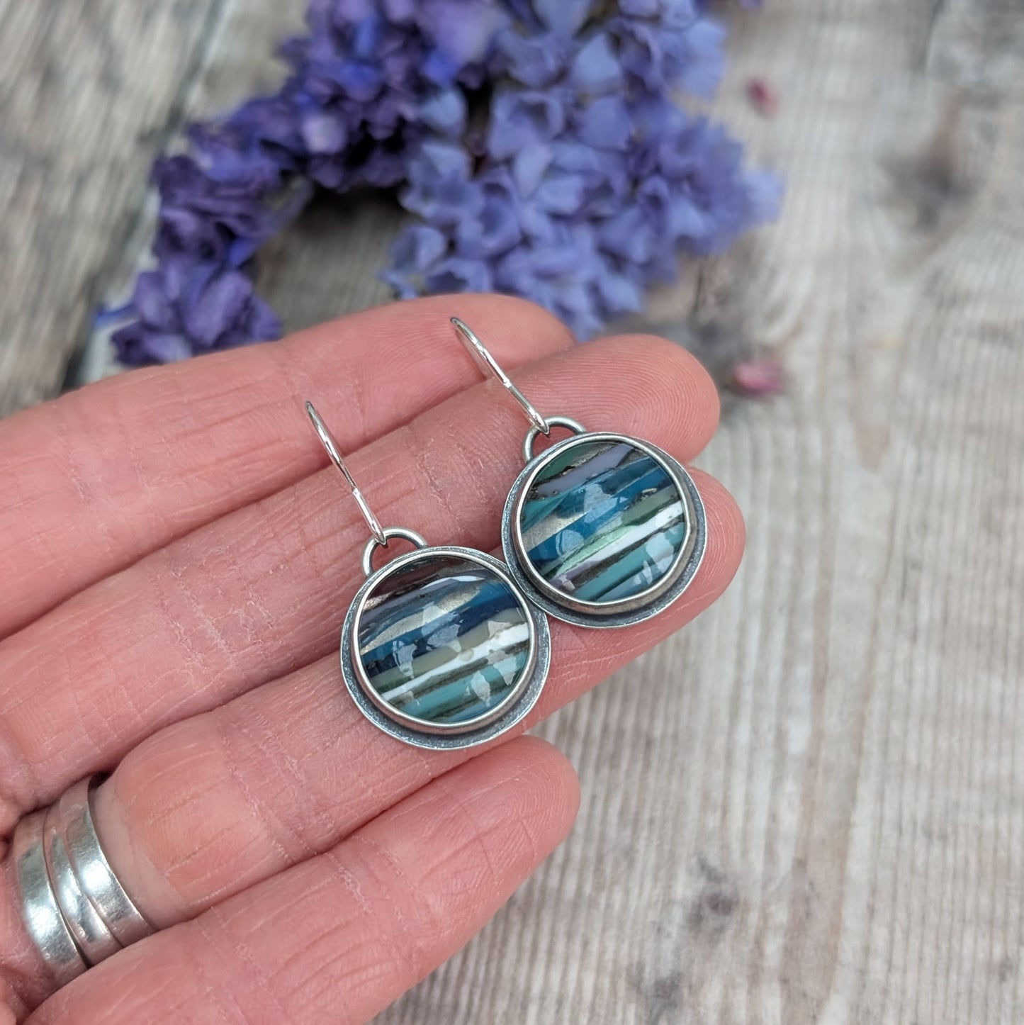 Each earring is a round surfite stone set in an oxidised silver bezel, suspended from silver earring wire. Surfite has random horizontal uneven stripes of lilac, blue, turquoise, green, white and black, gloss finish.