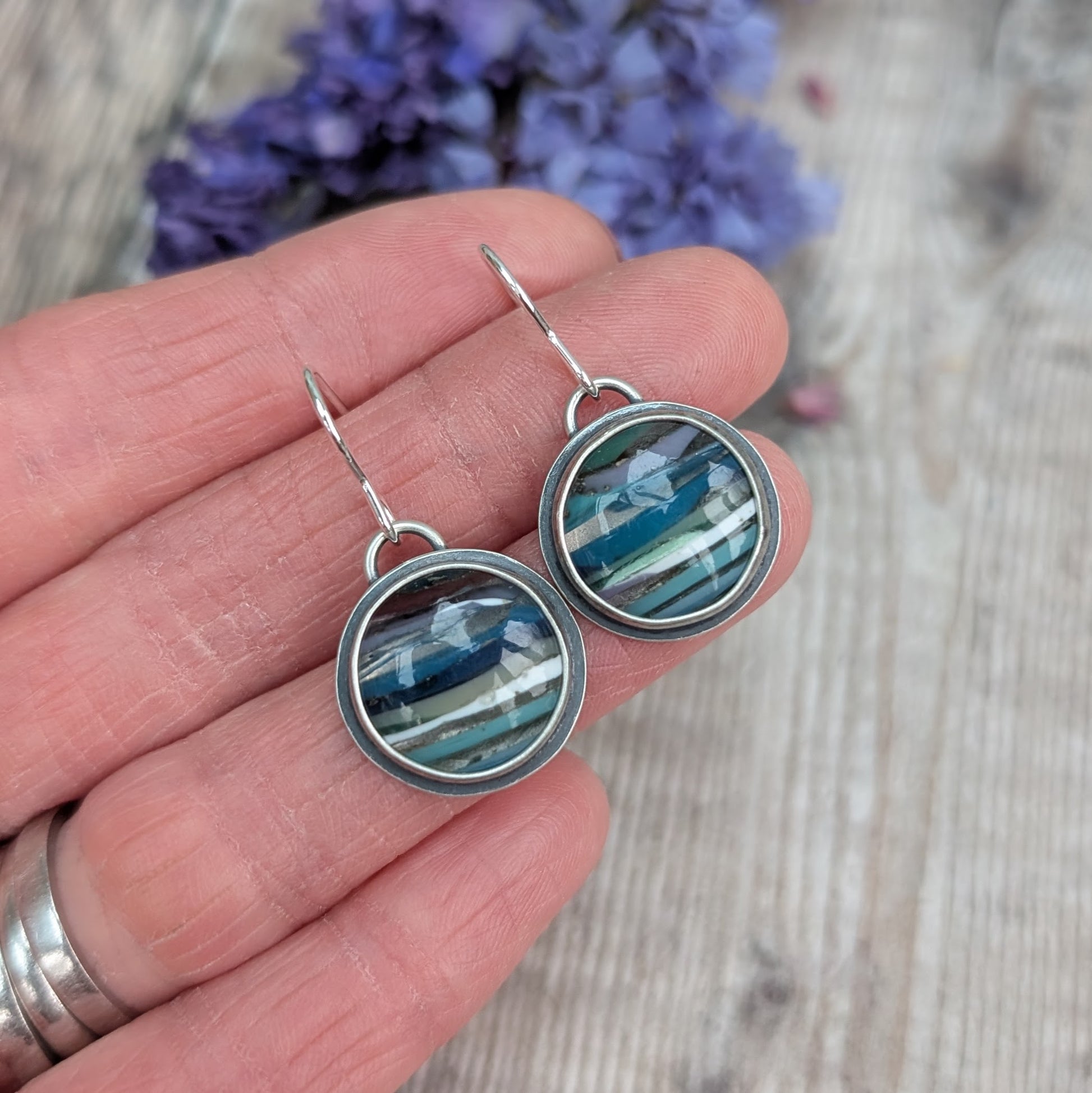 Each earring is a round surfite stone set in an oxidised silver bezel, suspended from silver earring wire. Surfite has random horizontal uneven stripes of lilac, blue, turquoise, green, white and black, gloss finish.