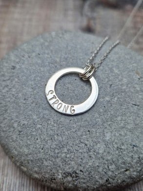 Sterling Silver ‘STRONG’ necklace. Silver disc with offset hole in centre, looks like a thick circle. Attached via two small silver hoops to silver chain. On thicker part of disc, inscription wording hammered into the silver reads STRONG in capitals. Disc measures approximately 20 mm in diameter. Chain length 16, 18 or 20 inches.