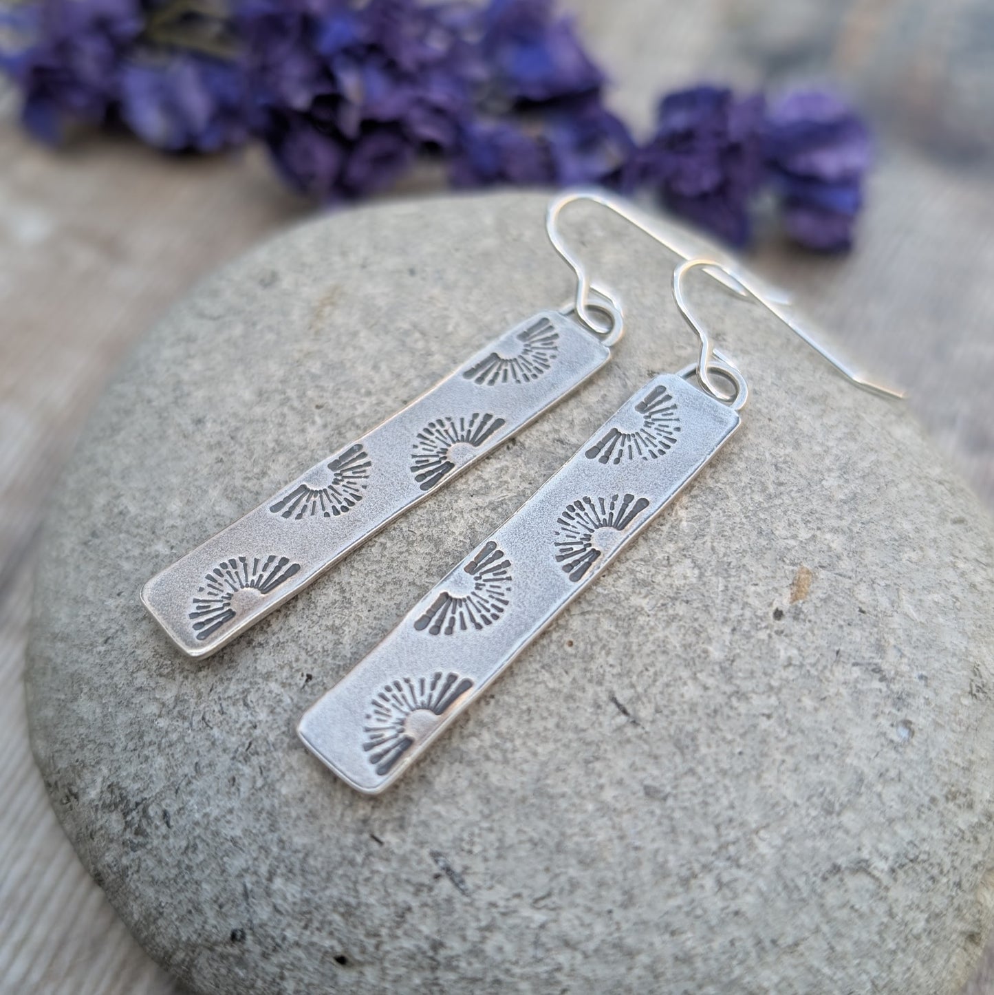 A pair of oxidised sterling silver earrings with a stamped sunburst design, displayed on a smooth stone with purple flowers in the background.