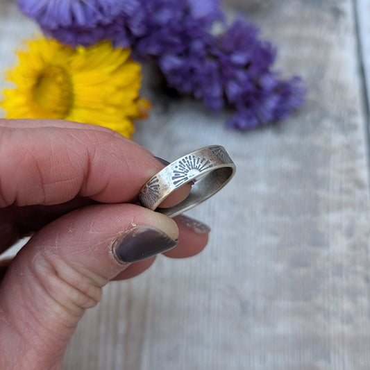 A detailed close-up of the sunburst stamped silver ring being held between two fingers, with a floral backdrop enhancing the handmade texture.