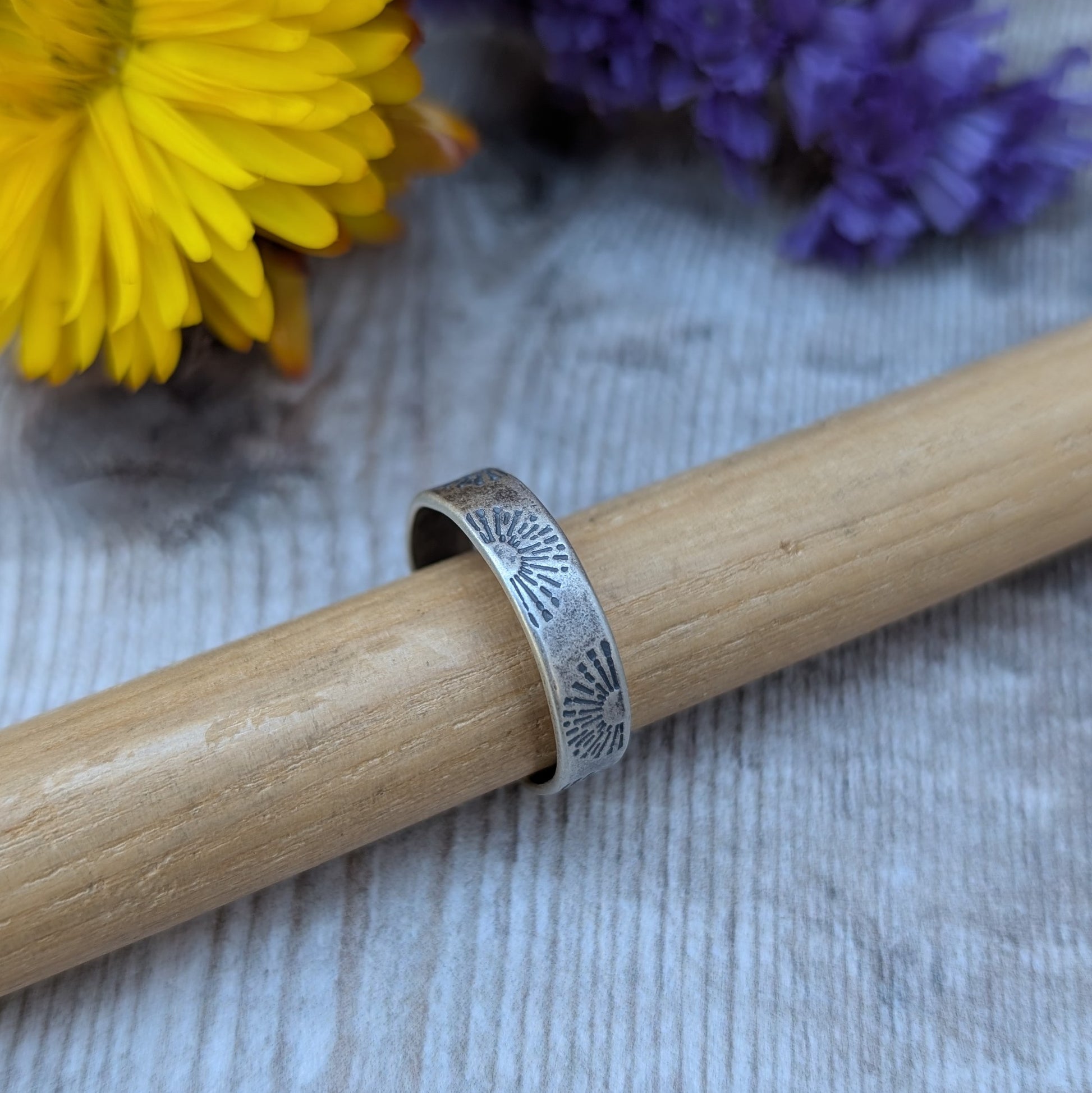 A close-up of an oxidised sterling silver ring with a sunburst stamped pattern, displayed on a wooden dowel with yellow and purple flowers in the background.