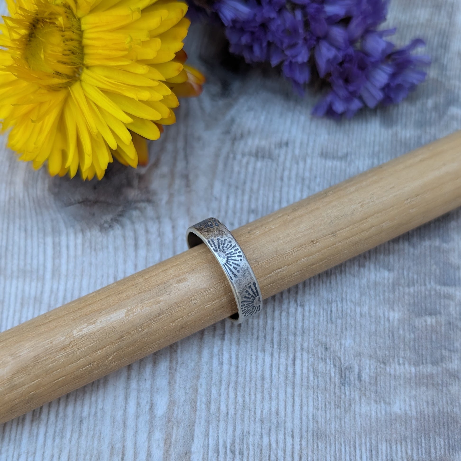 A close-up of an oxidised sterling silver ring with a sunburst stamped pattern, displayed on a wooden dowel with yellow and purple flowers in the background.