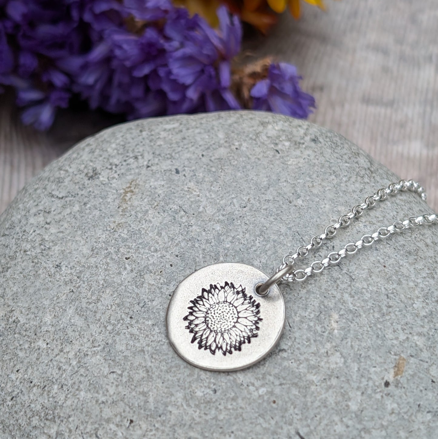 Close-up of a silver sunflower disc necklace lying on a smooth grey stone, with soft purple flowers in the background