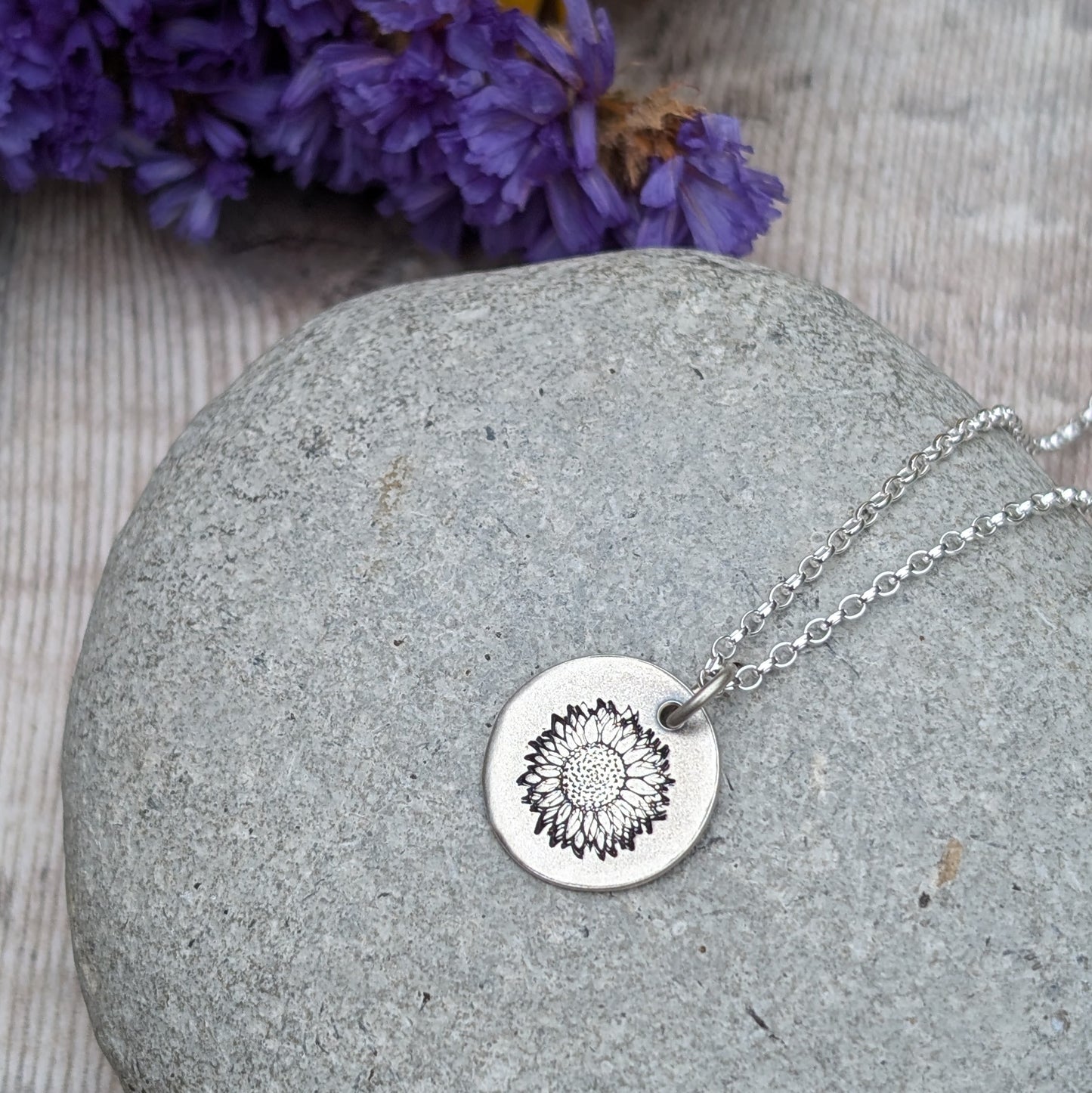 A silver sunflower disc necklace resting on a smooth grey stone, with purple flowers in the background