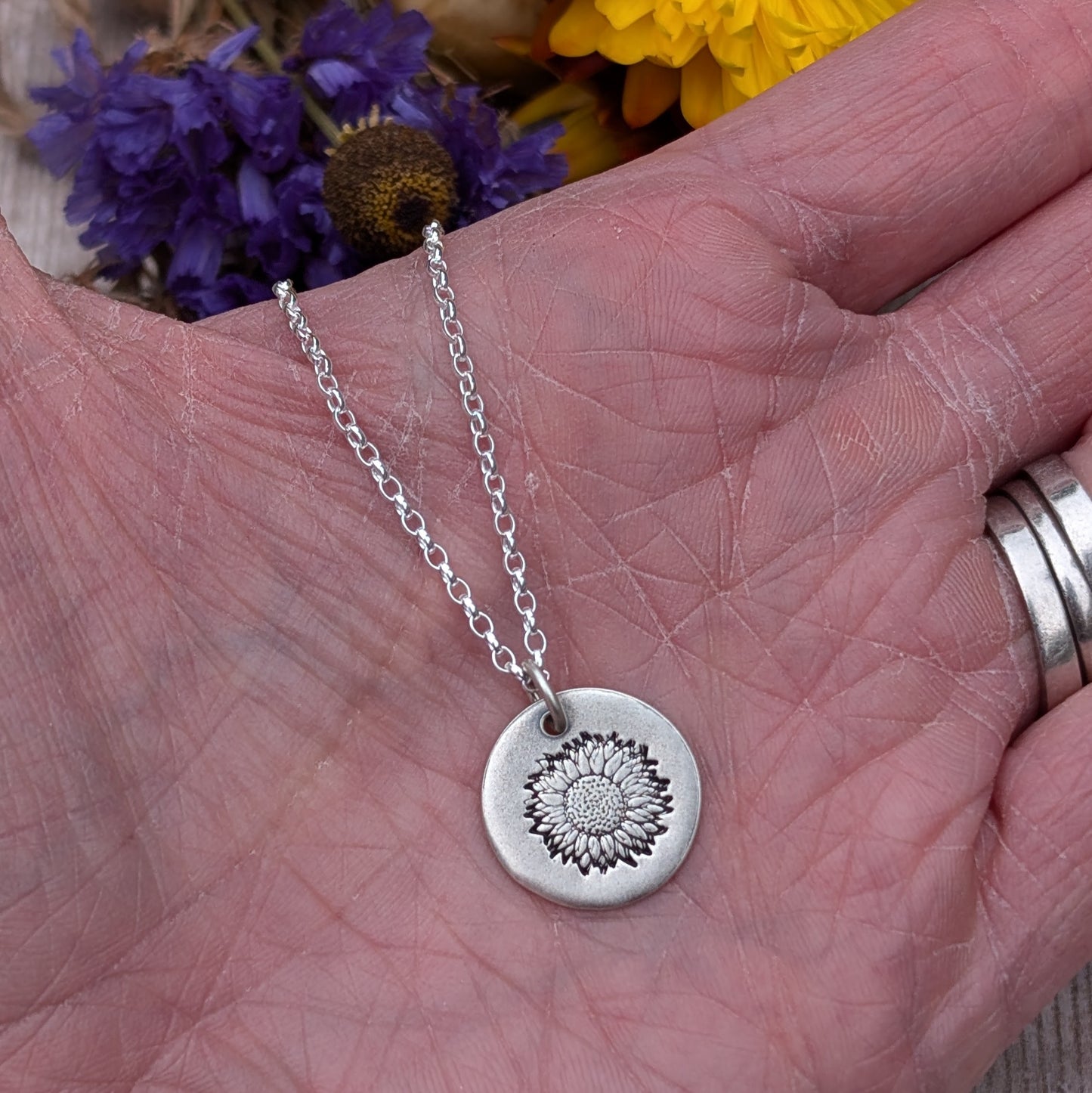 A silver sunflower disc necklace displayed on the palm of a hand, with dried yellow and purple flowers in the background.