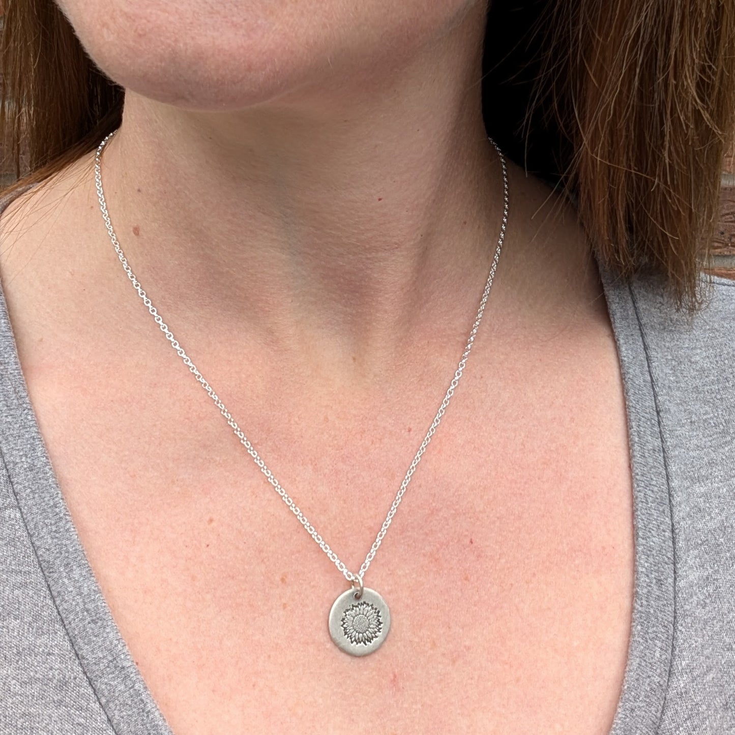 A silver sunflower disc necklace worn on a woman’s neck, shown against a grey top.