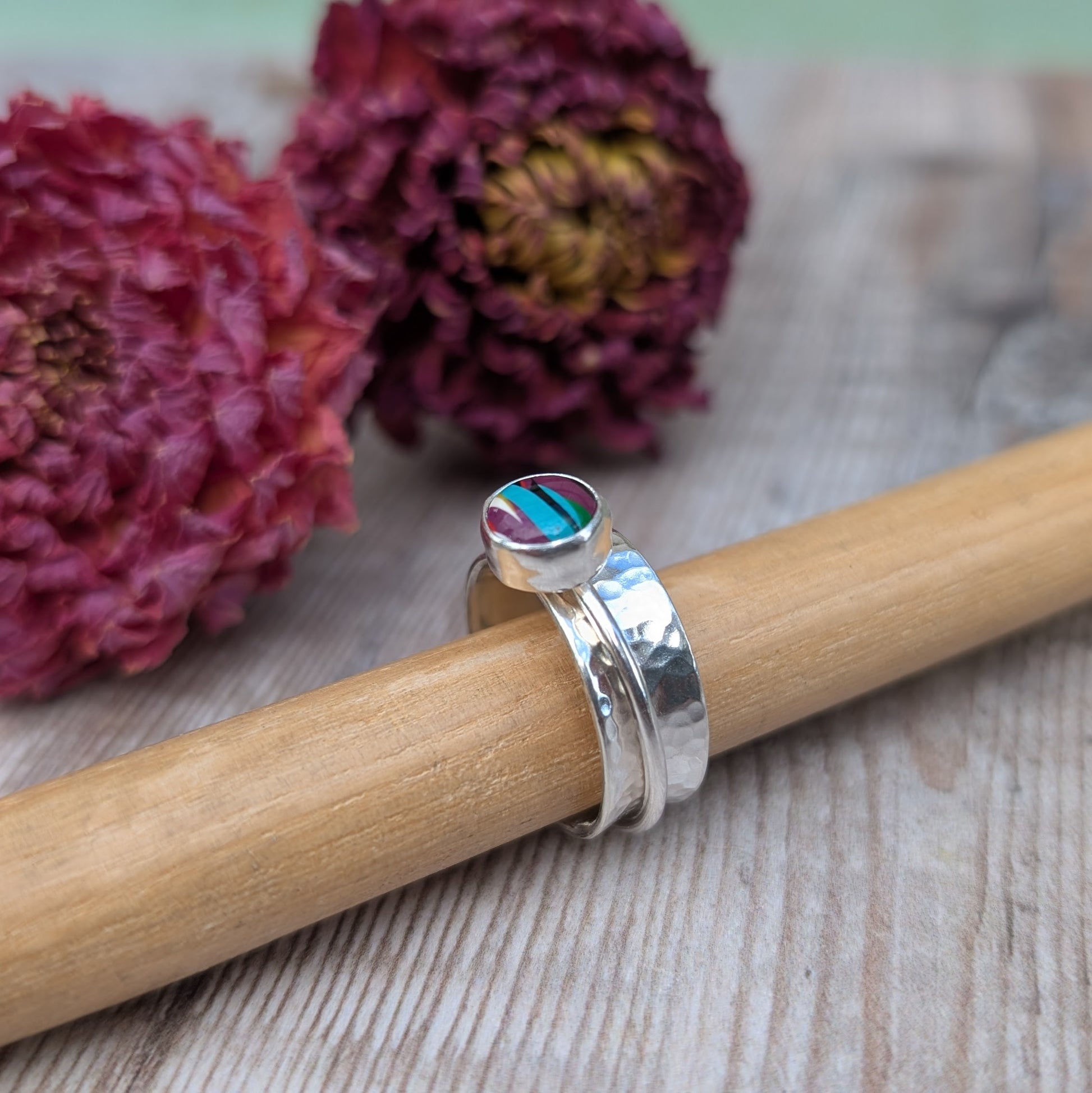 A handmade recycled sterling silver spinner ring featuring a bezel-set Surfite stone in shades of purple, teal, and yellow, displayed on a wooden ring mandrel with dried flowers in the background.