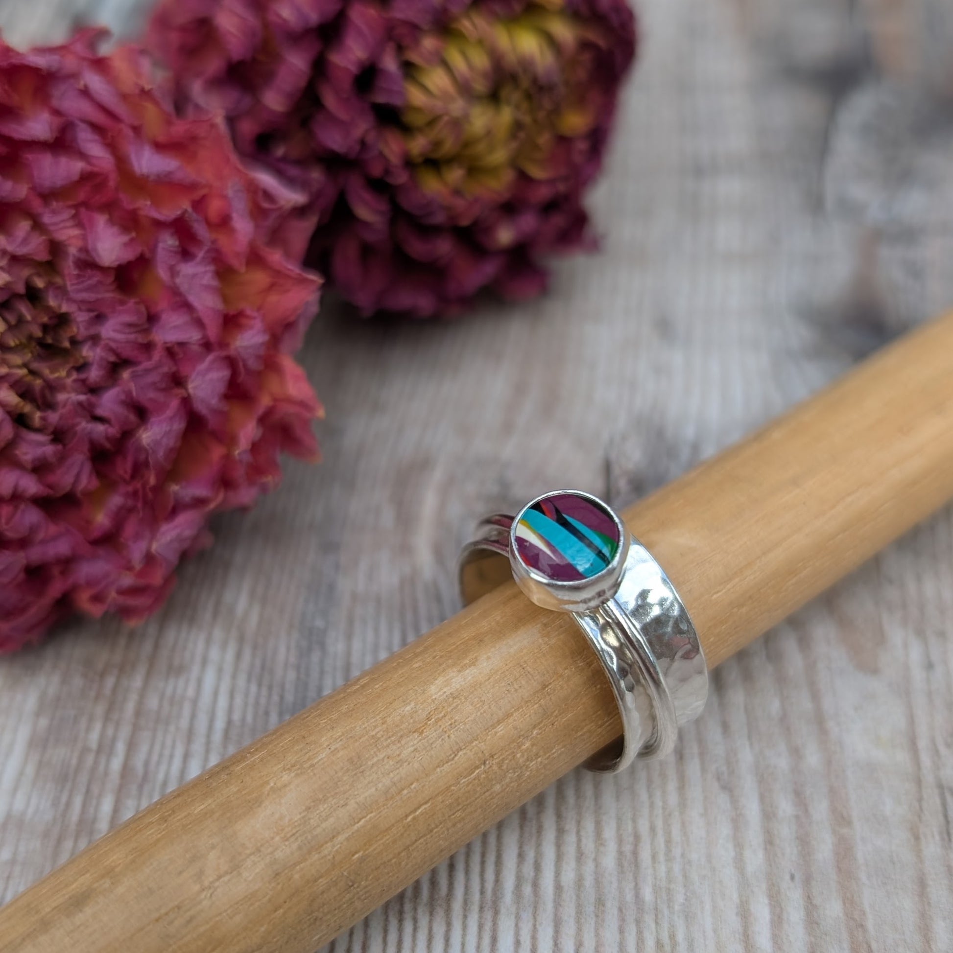 A handmade recycled sterling silver spinner ring featuring a bezel-set Surfite stone in shades of purple, teal, and yellow, displayed on a wooden ring mandrel with dried flowers in the background.