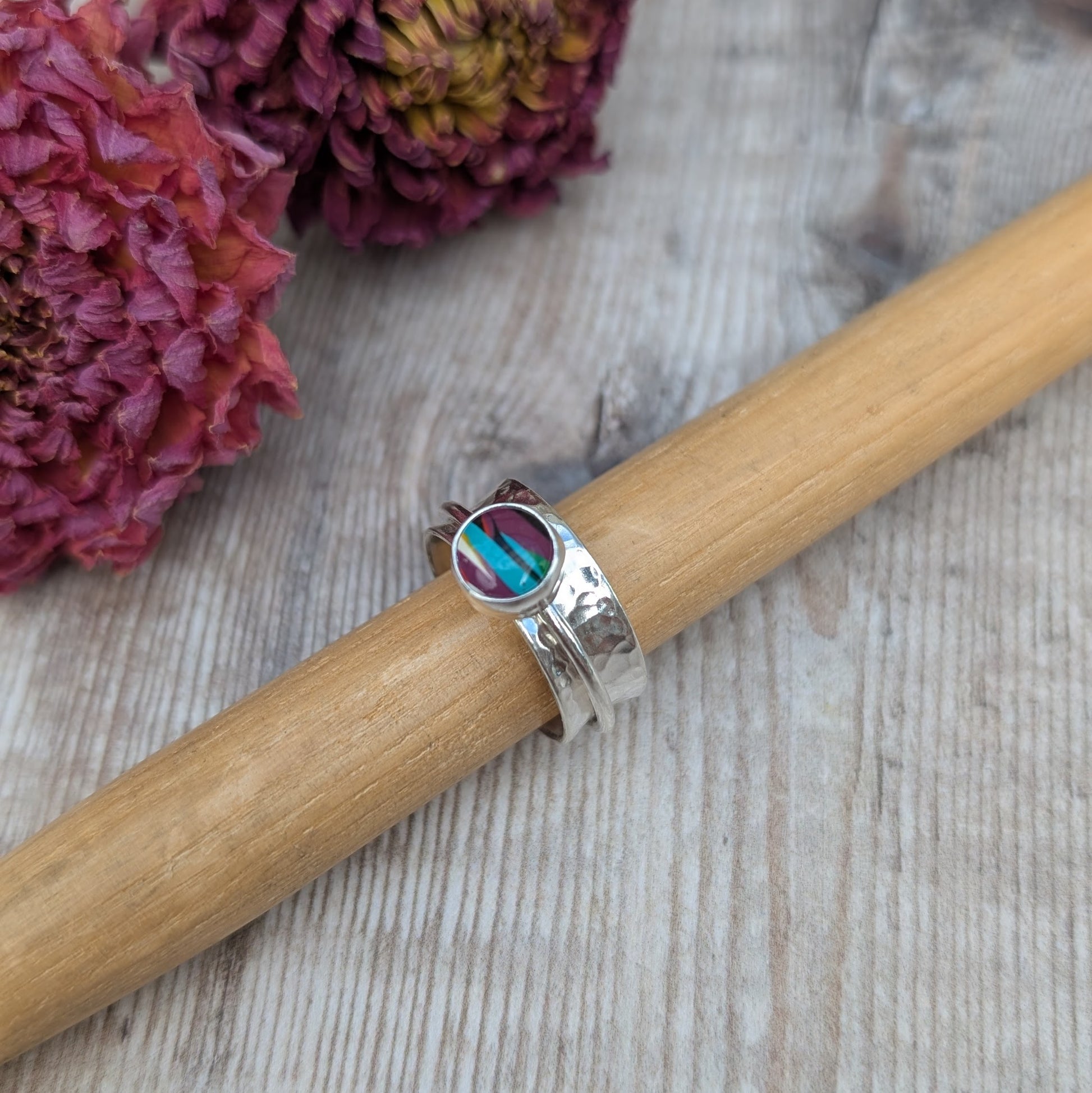 A handmade recycled sterling silver spinner ring featuring a bezel-set Surfite stone in shades of purple, teal, and yellow, displayed on a wooden ring mandrel with dried flowers in the background.