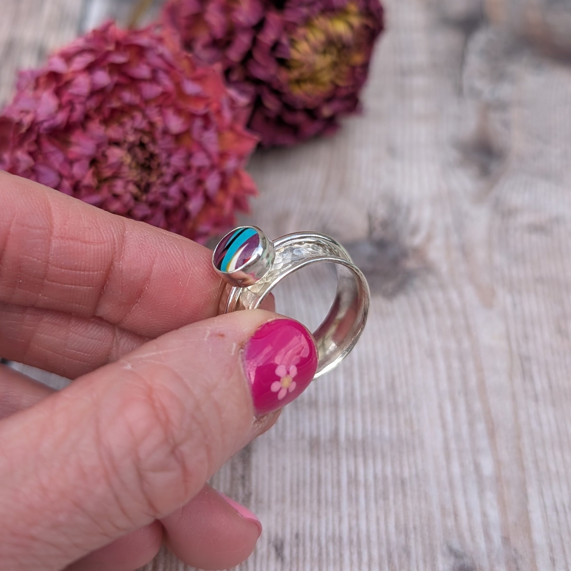 A person holding the Surfite spinner ring between their fingers, highlighting the textured silver band and the unique layered colours of the stone, with a rustic wooden surface and dried flowers in the background.