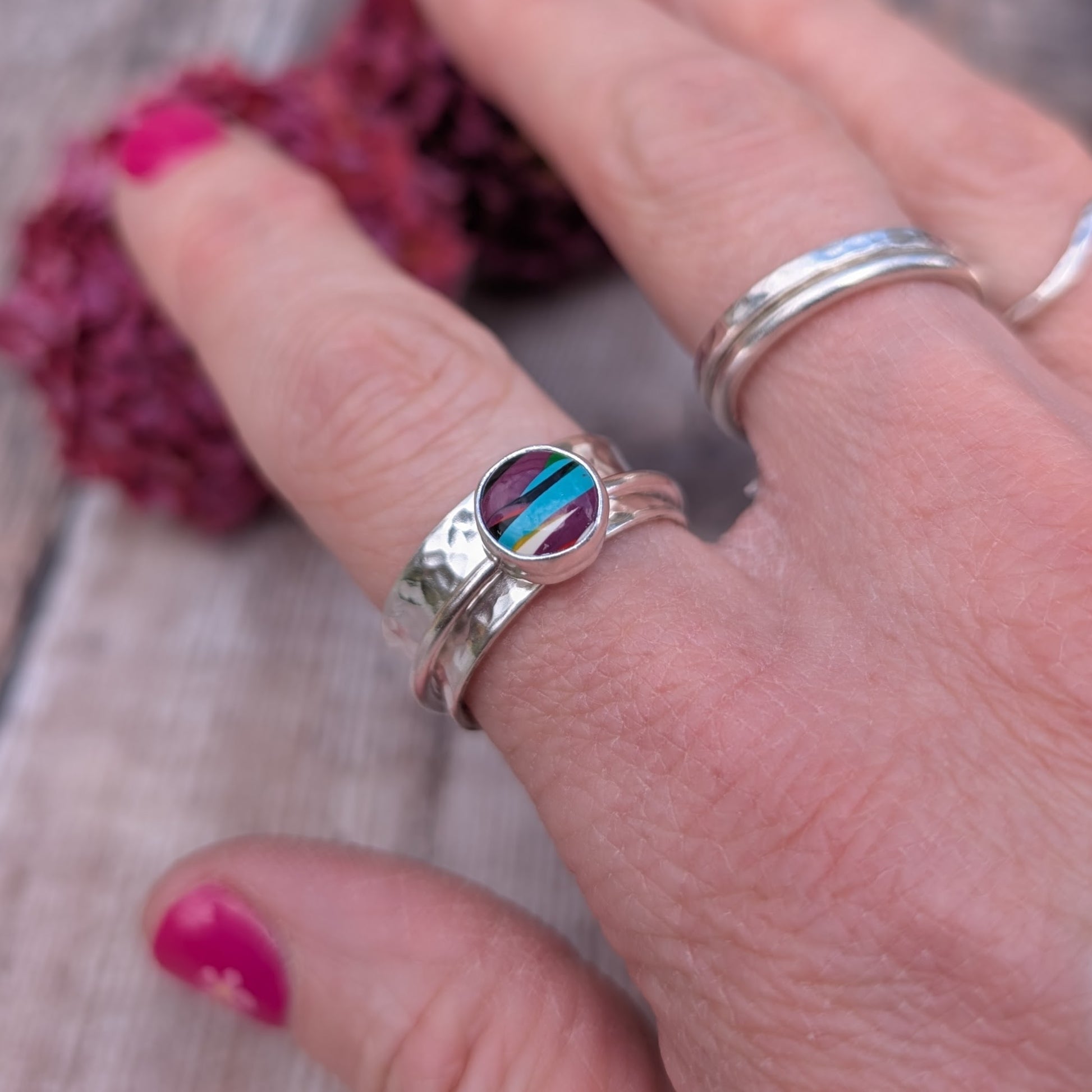 A close-up of a hand wearing the Surfite spinner ring, showing the hammered silver band and the colourful Surfite stone, with additional silver rings worn on other fingers.
