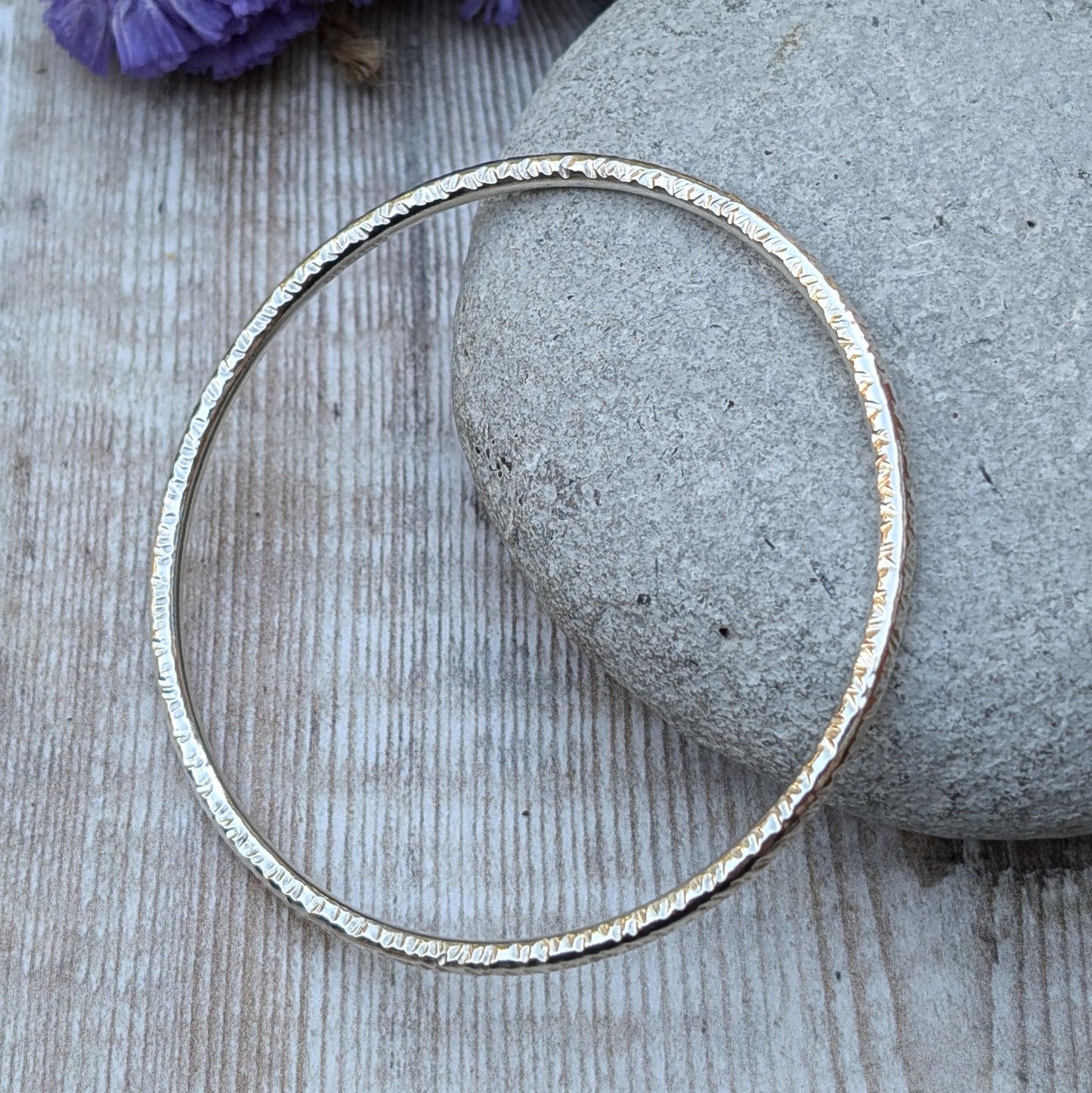 A handmade sterling silver bangle with a hammered texture is displayed resting on a smooth grey stone. The subtle light reflections highlight the intricate hammered details of the bangle, with soft purple and yellow flowers in the background.
