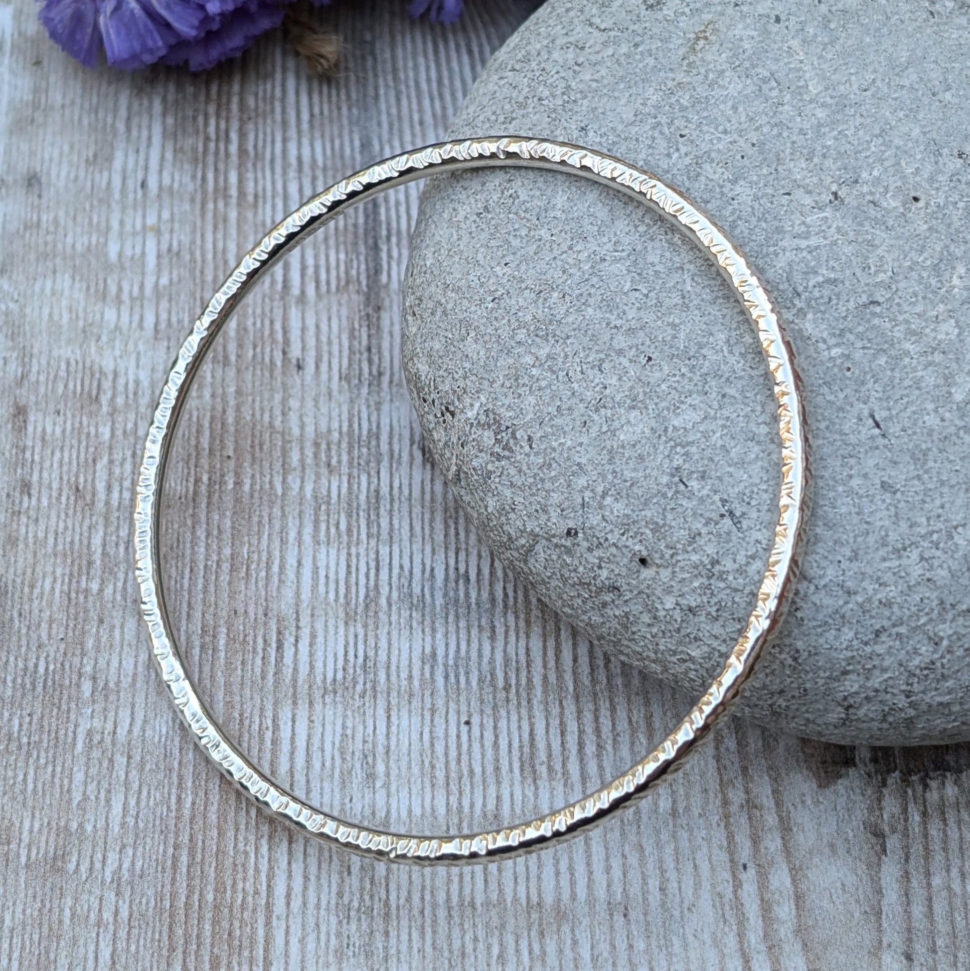 A handmade sterling silver bangle with a hammered texture is displayed resting on a smooth grey stone. The subtle light reflections highlight the intricate hammered details of the bangle, with soft purple and yellow flowers in the background.