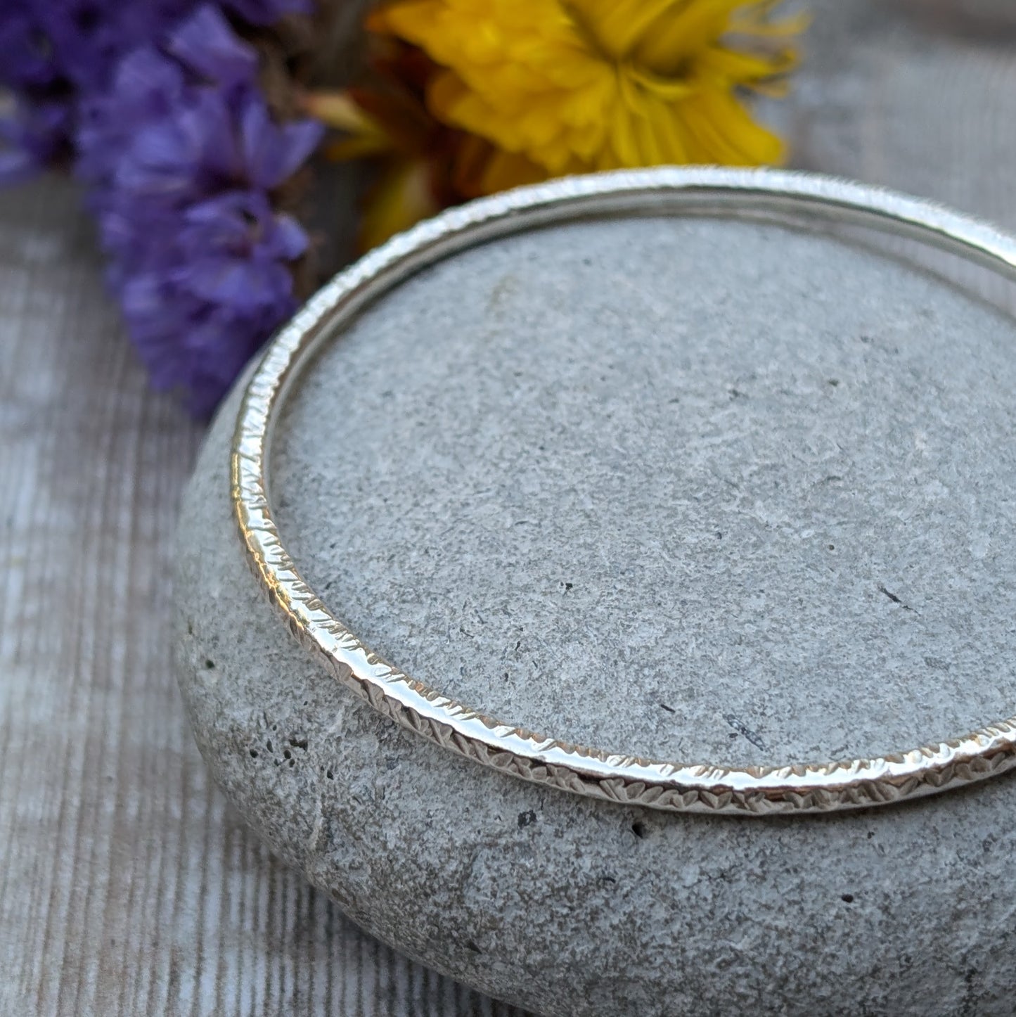 A handmade sterling silver bangle with a hammered texture is displayed resting on a smooth grey stone. The subtle light reflections highlight the intricate hammered details of the bangle, with soft purple and yellow flowers in the background.