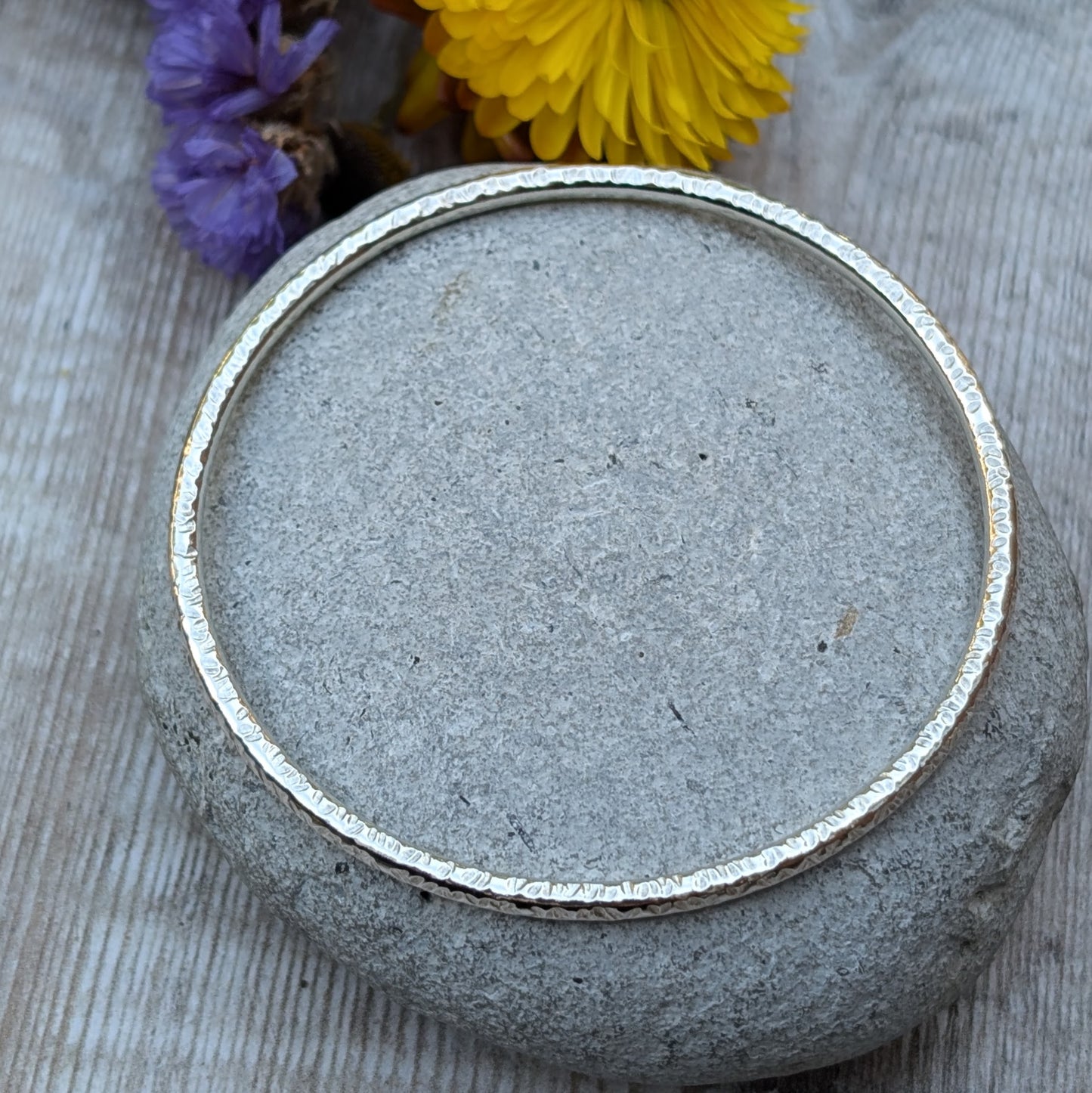 A handmade sterling silver bangle with a hammered texture is displayed resting on a smooth grey stone. The subtle light reflections highlight the intricate hammered details of the bangle, with soft purple and yellow flowers in the background.