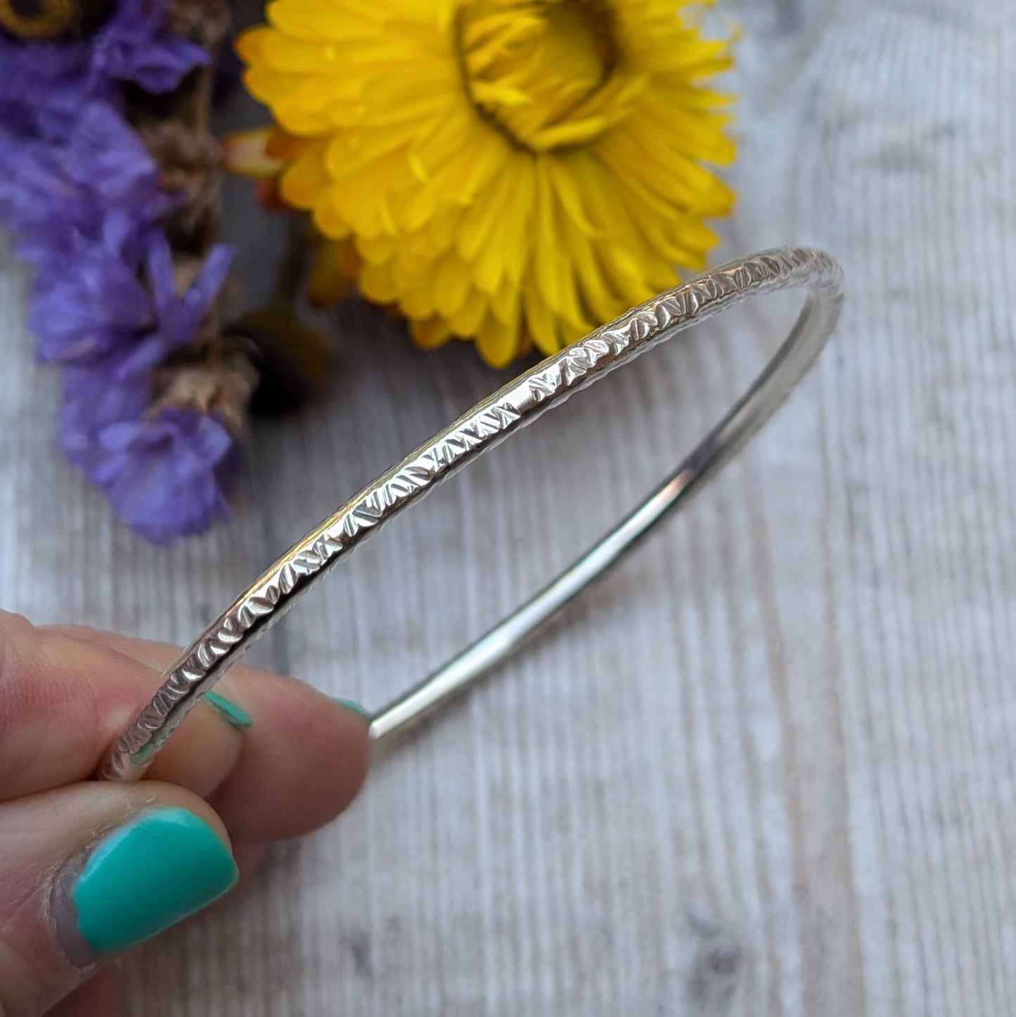 A close-up of a handmade sterling silver bangle with a hammered pattern, held gently between fingers. The textured surface catches the light, while vibrant yellow and purple flowers serve as a colorful backdrop.