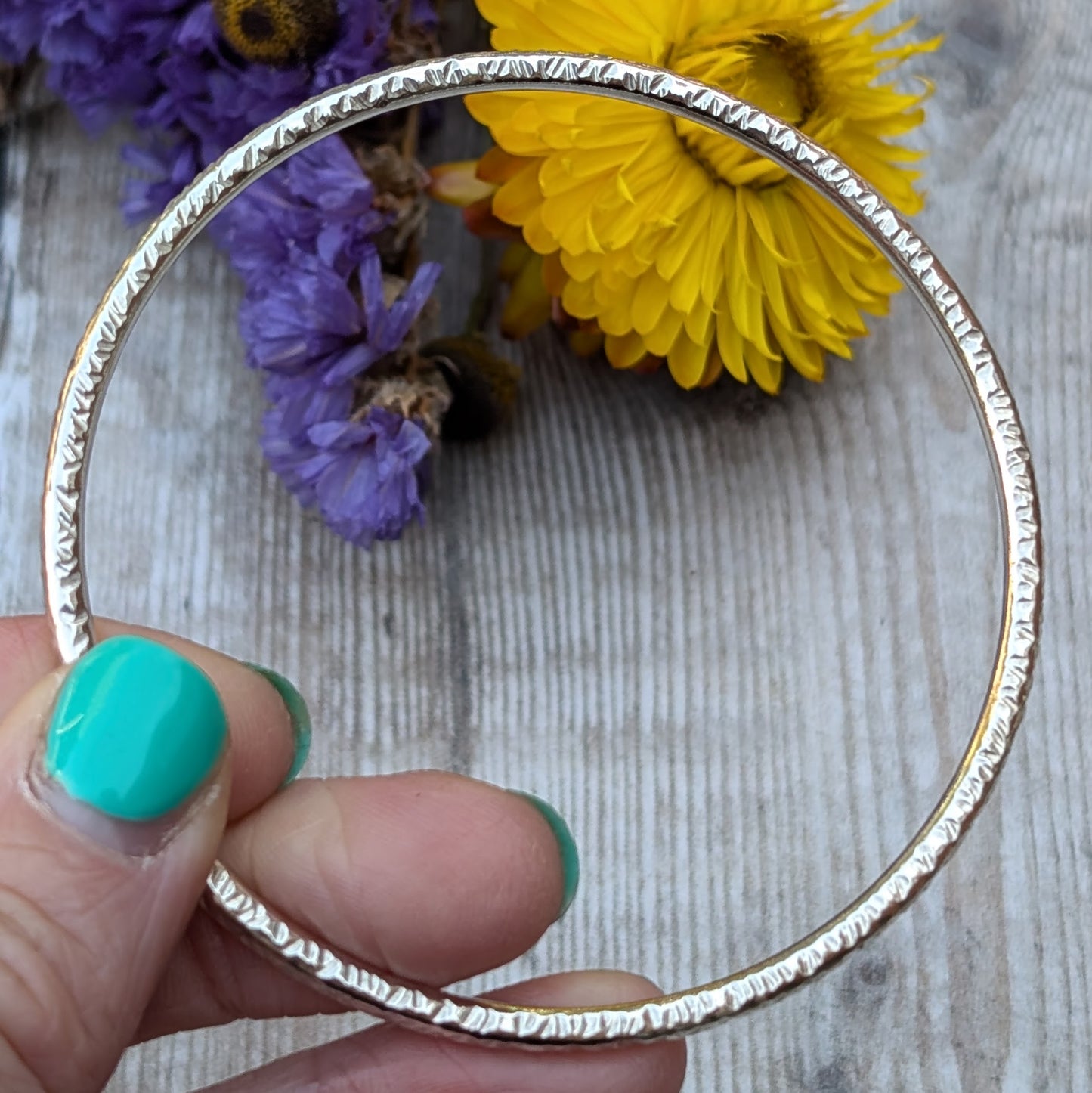 A close-up of a handmade sterling silver bangle with a hammered pattern, held gently between fingers. The textured surface catches the light, while vibrant yellow and purple flowers serve as a colorful backdrop.