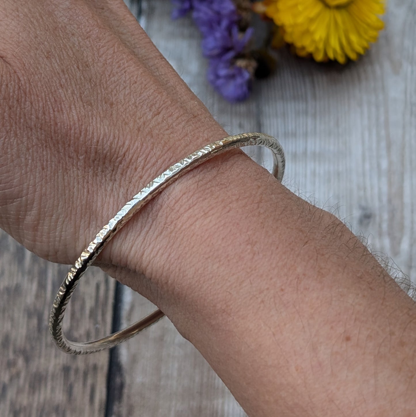 A handmade sterling silver bangle with a hammered finish is shown on a wrist, reflecting light and displaying the fine details of the texture. The neutral tone of the skin contrasts with the shiny silver, and a rustic wooden surface is visible in the background.