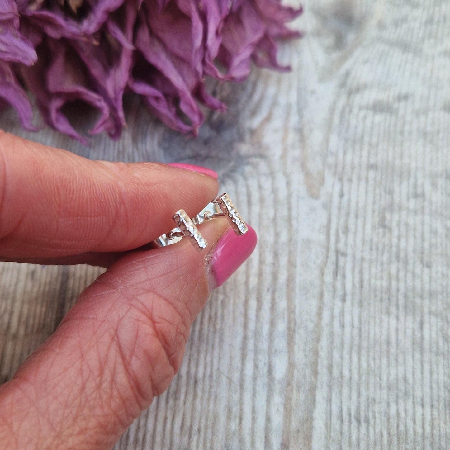 Close-up of a hand holding tiny sterling silver stud earrings with a hammered twig design, showcasing their size and texture against a rustic background