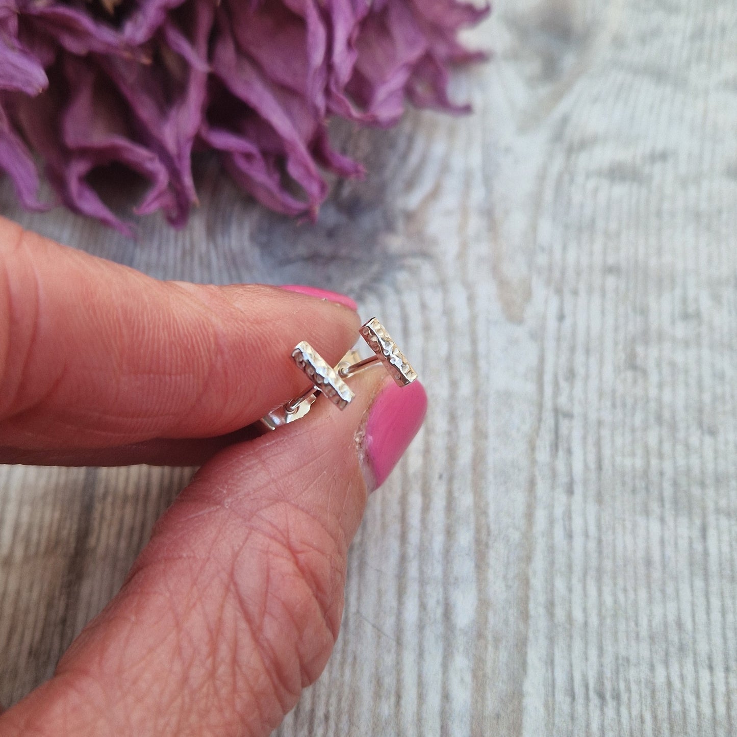 Close-up of a hand holding tiny sterling silver stud earrings with a hammered twig design, showcasing their size and texture against a rustic background