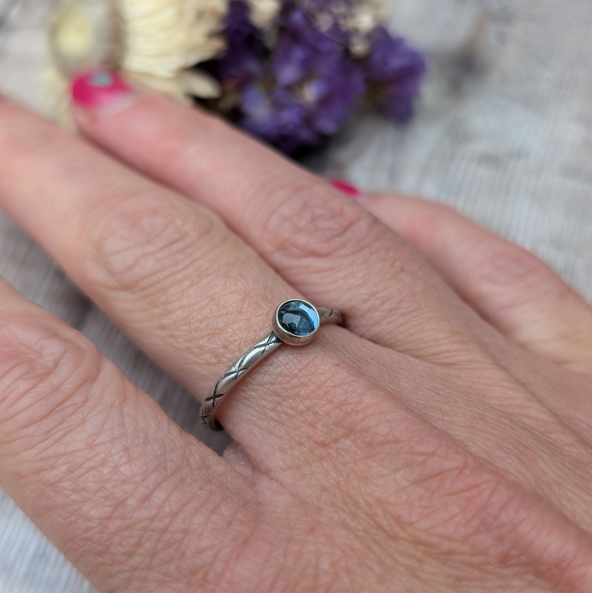 Slim silver band with cross stamped pattern. A blue topaz round stone is mounted on the band in an oxidised sterling silver bezel. Rings can be stacked above or below this piece.