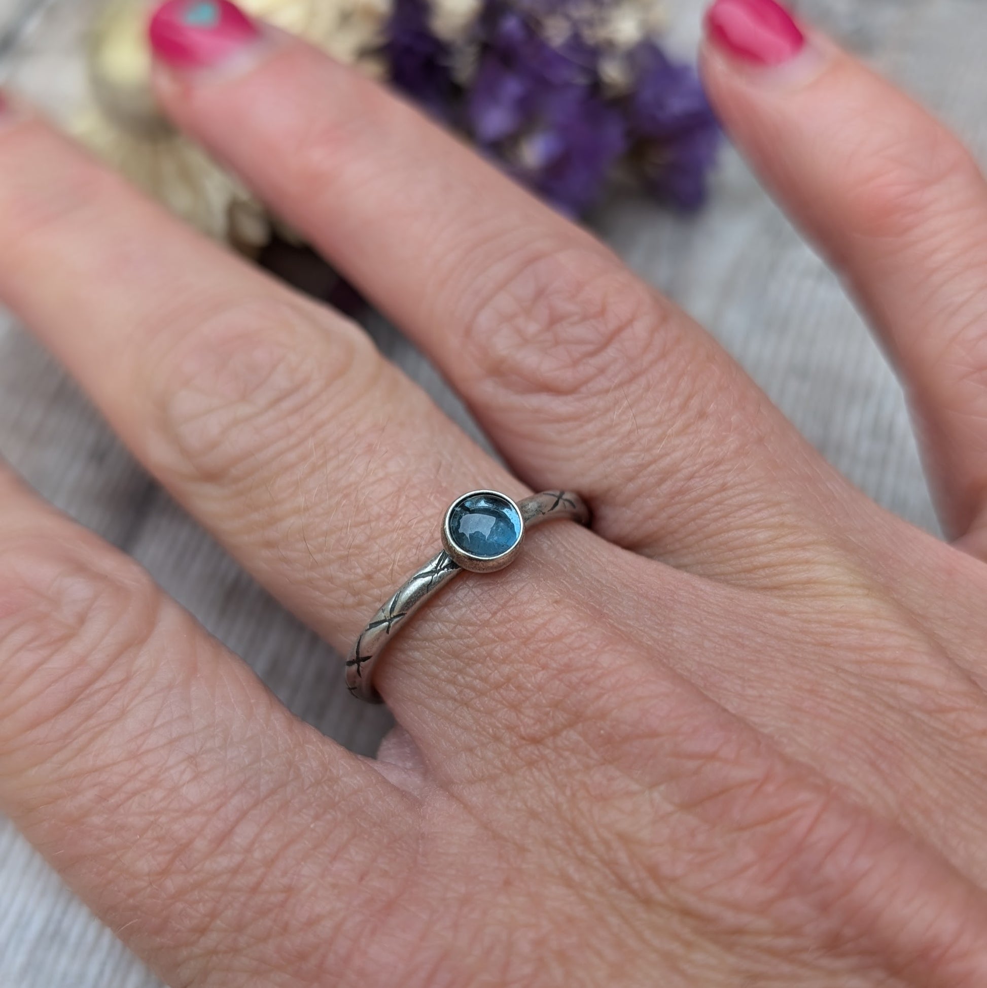 Slim silver band with cross stamped pattern. A blue topaz round stone is mounted on the band in an oxidised sterling silver bezel. Rings can be stacked above or below this piece.
