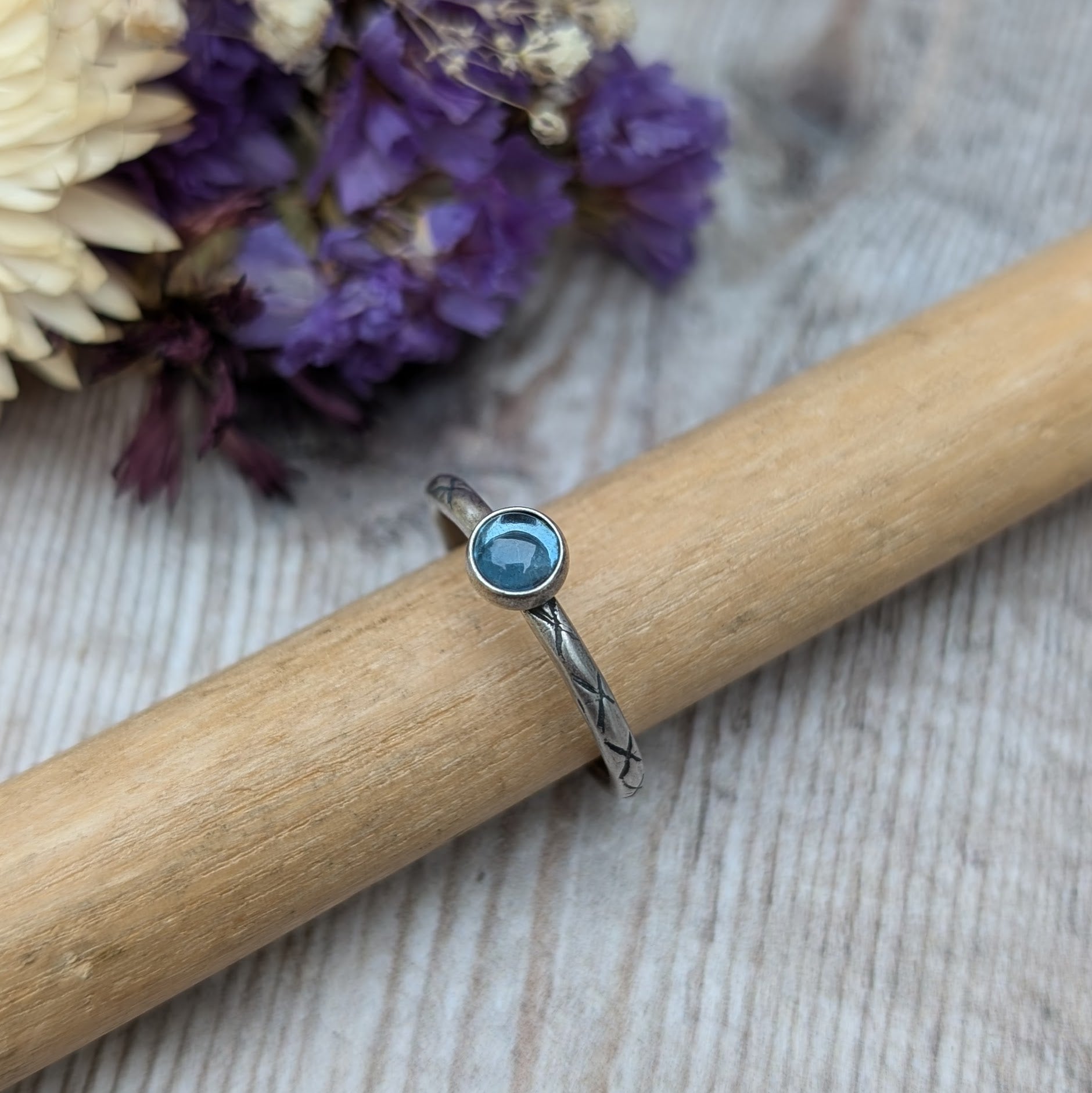 Slim silver band with cross stamped pattern. A blue topaz round stone is mounted on the band in an oxidised sterling silver bezel. Rings can be stacked above or below this piece.