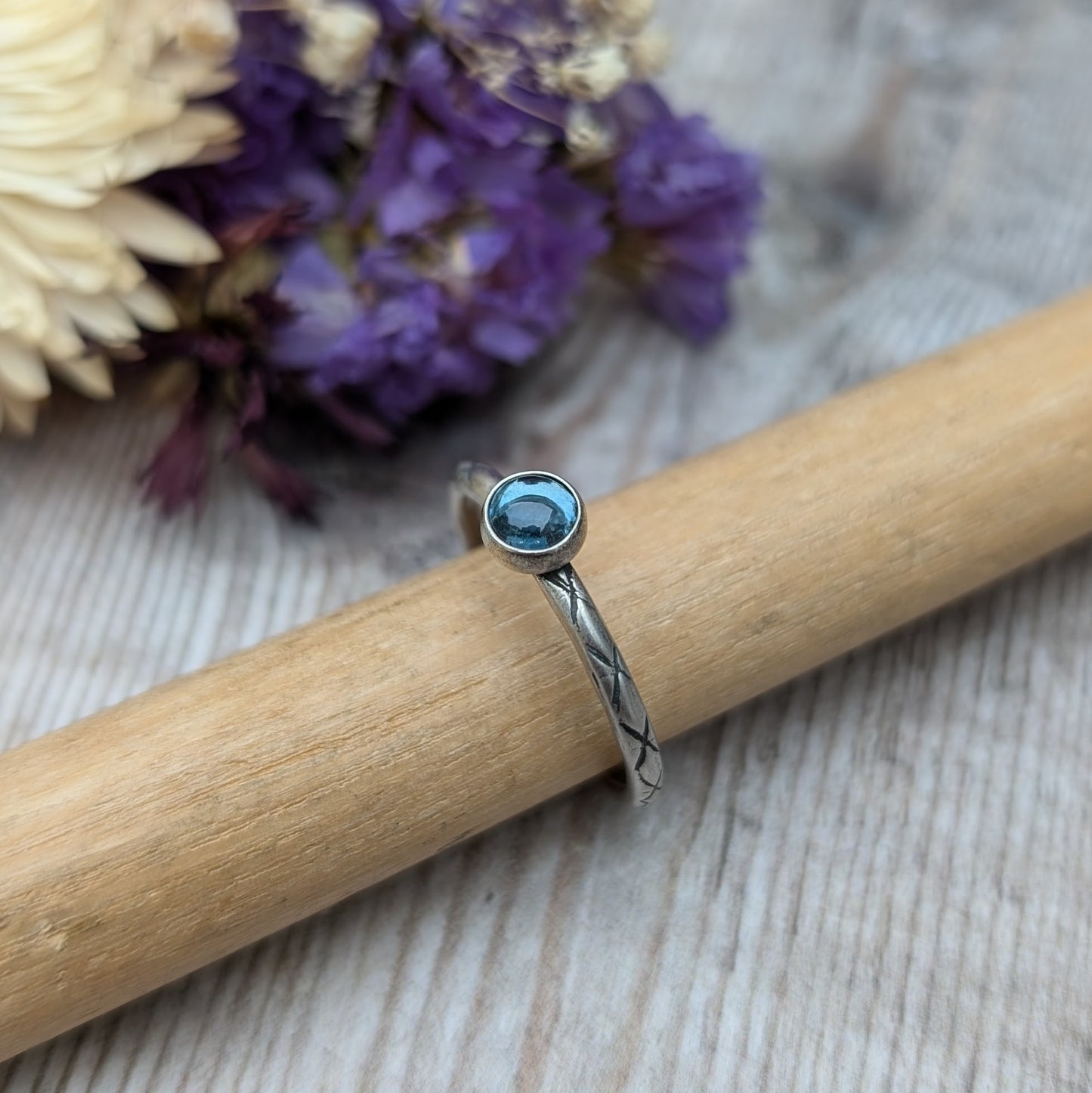 Slim silver band with cross stamped pattern. A blue topaz round stone is mounted on the band in an oxidised sterling silver bezel. Rings can be stacked above or below this piece.