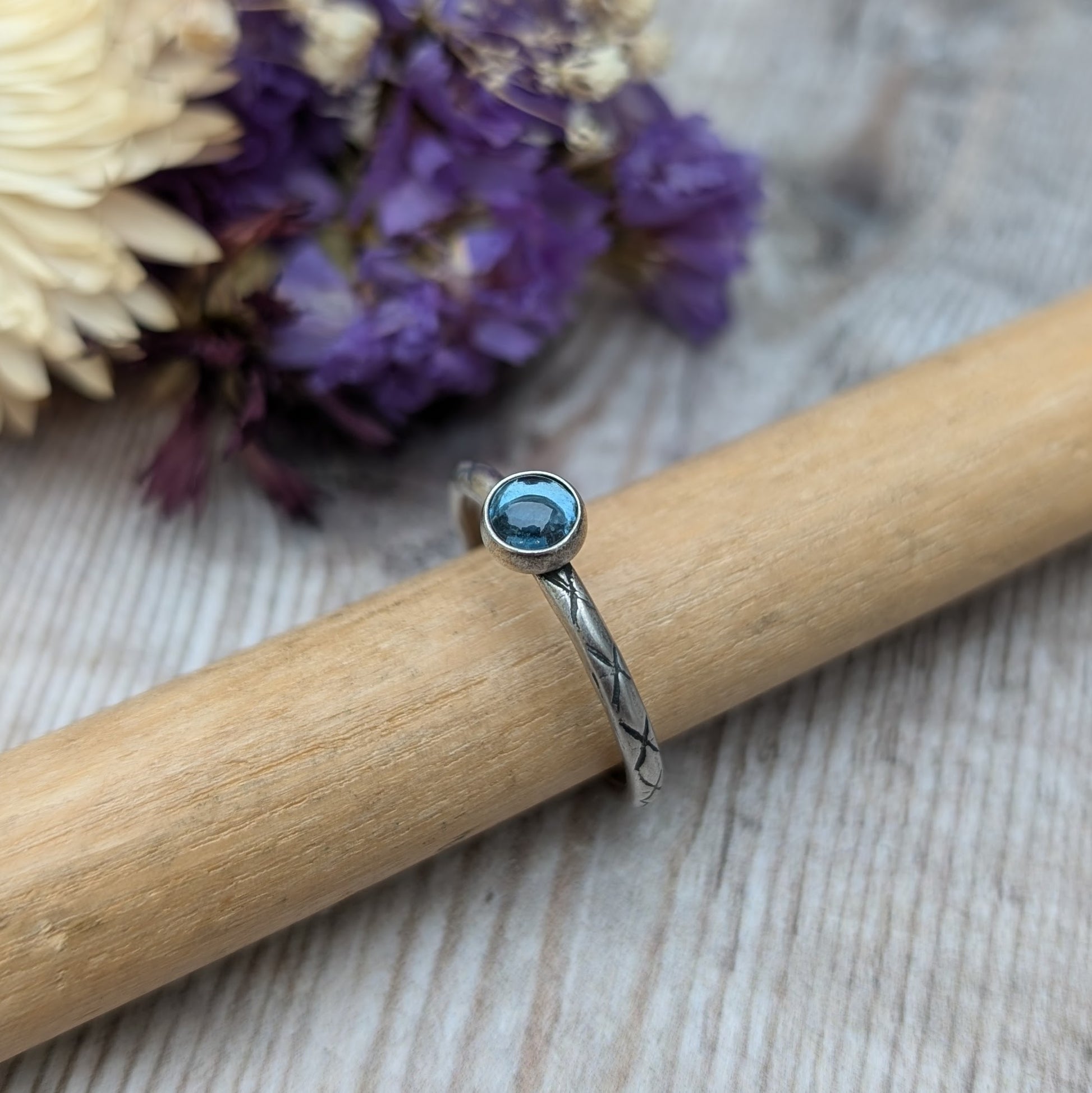 Slim silver band with cross stamped pattern. A blue topaz round stone is mounted on the band in an oxidised sterling silver bezel. Rings can be stacked above or below this piece.