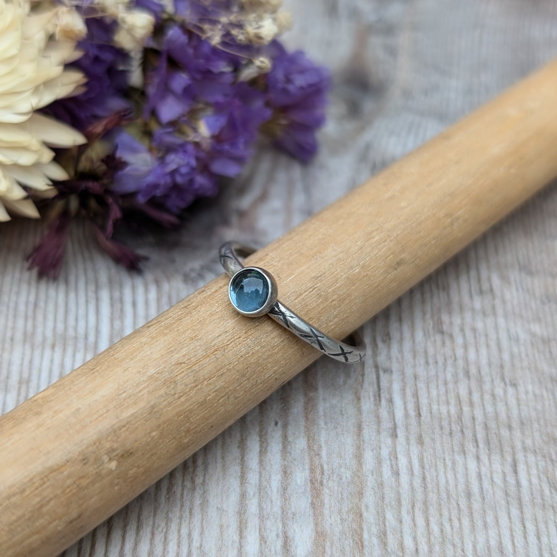 Slim silver band with cross stamped pattern. A blue topaz round stone is mounted on the band in an oxidised sterling silver bezel. Rings can be stacked above or below this piece.