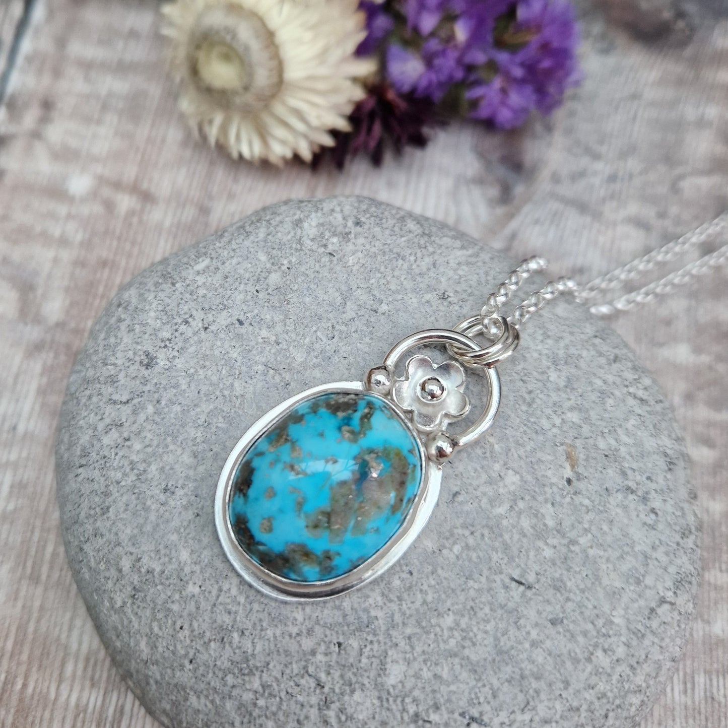 Turquoise oval gemstone pendant set in simple Sterling Silver bezel surround. Fused to the top of the bezel setting is a silver half circle with a five petaled flower in the middle, attached to silver chain via three small silver circles. Gemstone is predominantly turquoise blue with dark, almost black plume formations.