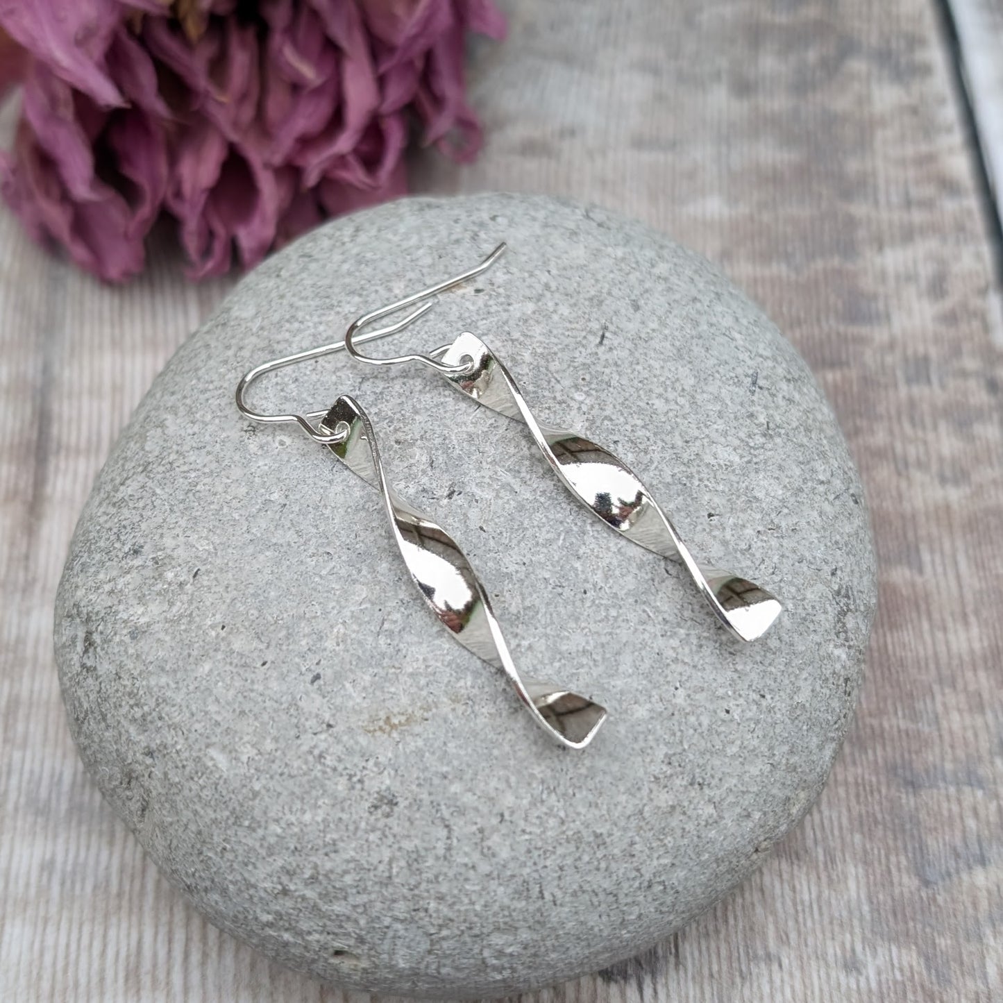 Pair of long, twisted silver earrings with a polished finish displayed on a smooth grey stone, with dried purple flowers blurred in the background.