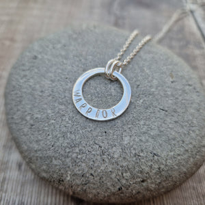 Sterling Silver ‘WARRIOR’ necklace. Silver disc with offset hole in centre, looks like a thick circle. Attached via two small silver hoops to silver chain. On thicker part of disc, inscription wording hammered into the silver reads WARRIOR in capitals. Disc measures approximately 20 mm in diameter. Chain length 16, 18 or 20 inches.