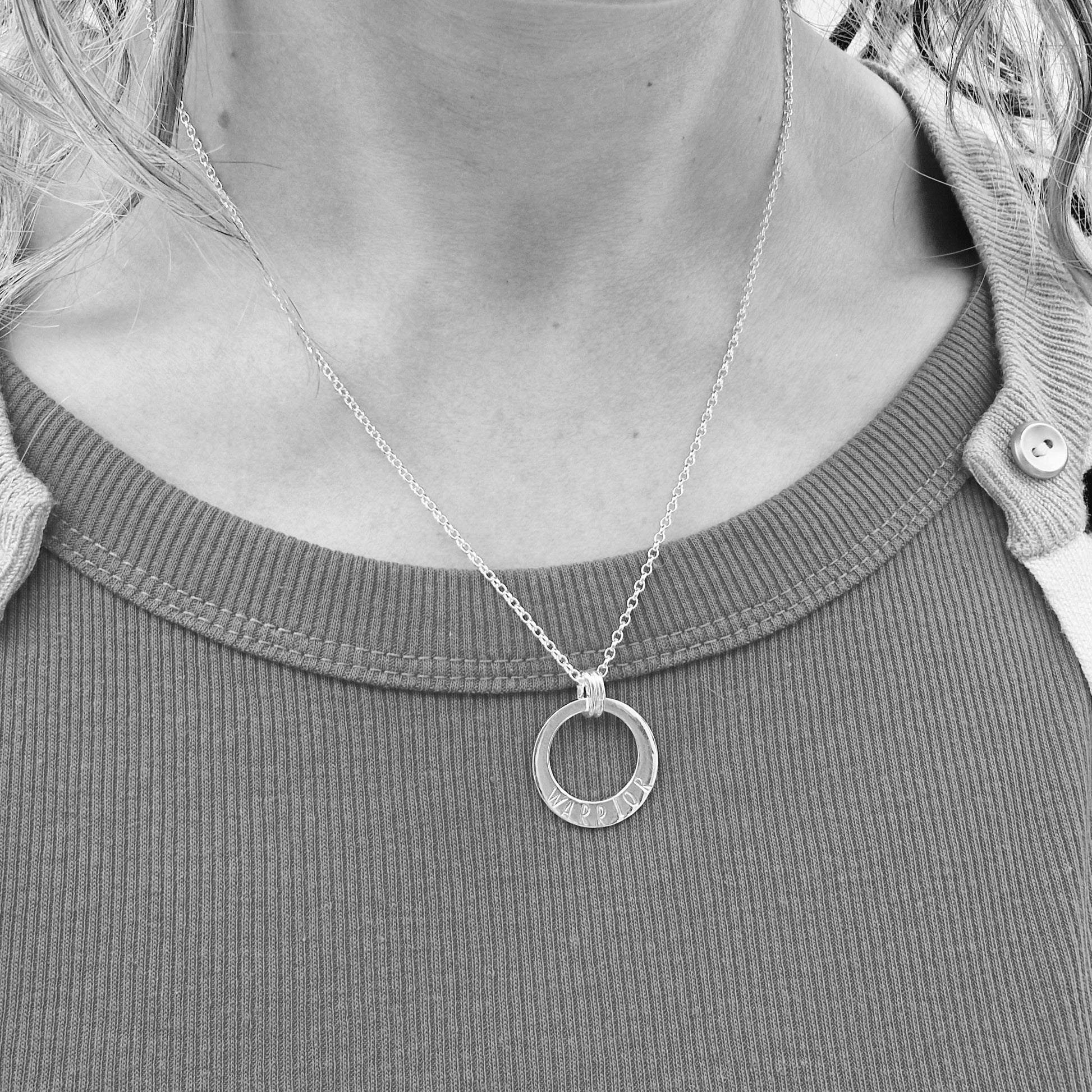 A black and white photo of a person wearing the silver "WARRIOR" circle necklace. The pendant hangs just below the collarbone on a fine silver chain, styled with a ribbed top and a cardigan draped over the shoulders.