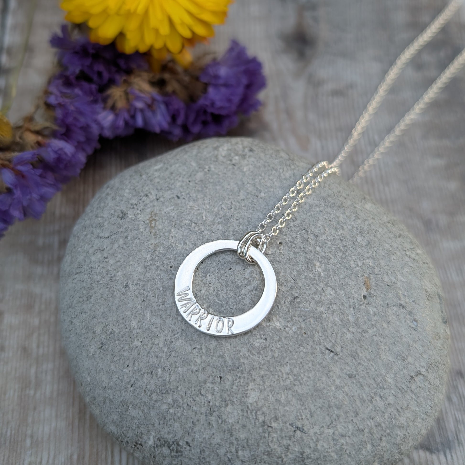A silver circle necklace with the word "WARRIOR" hand-stamped on the lower part of the pendant, resting on a smooth grey stone. The chain is draped over the stone, with a blurred background featuring dried purple flowers.