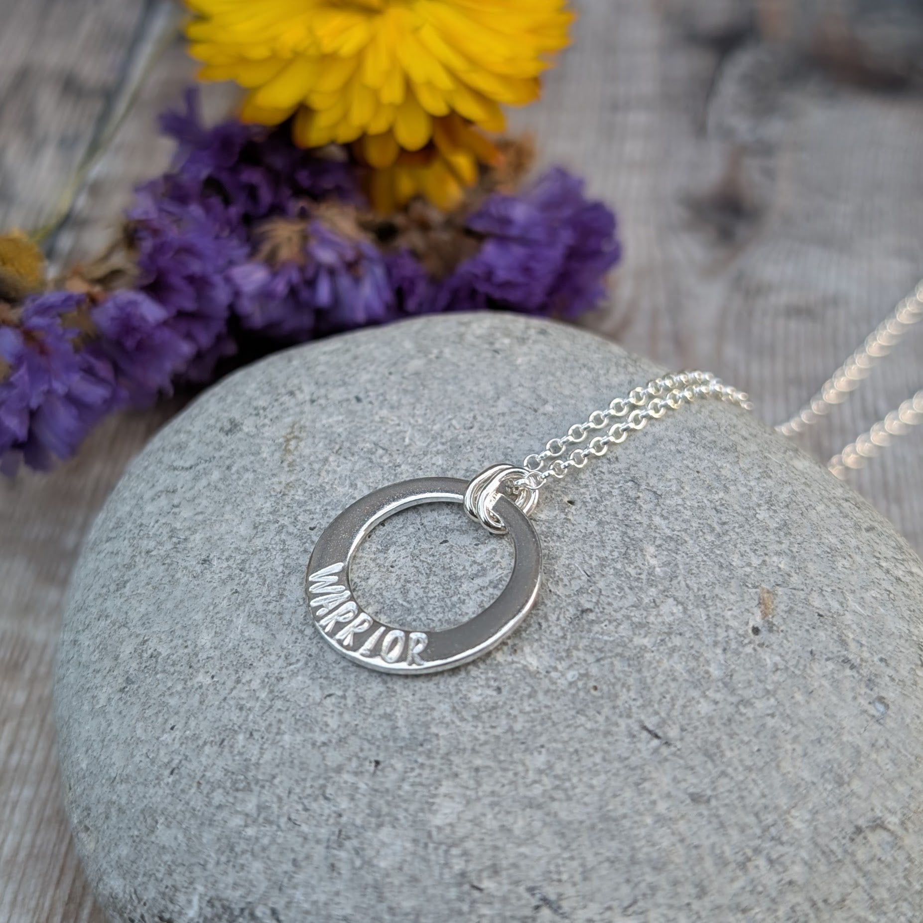 A silver circle necklace with the word "WARRIOR" hand-stamped on the lower part of the pendant, resting on a smooth grey stone. The chain is draped over the stone, with a blurred background featuring dried purple flowers.
