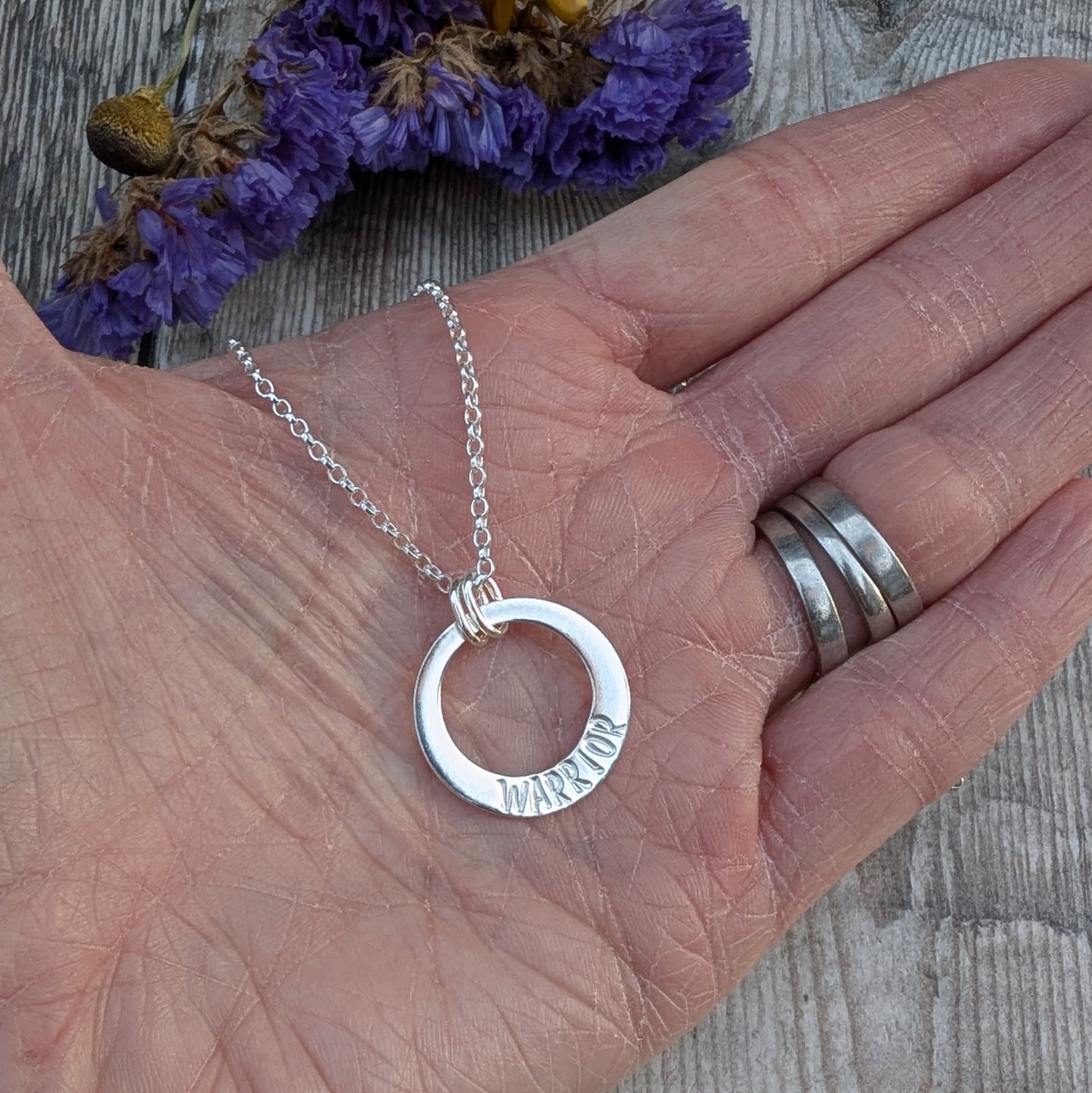 A close-up of a hand holding a silver circle necklace with the word "WARRIOR" hand-stamped on it. The pendant hangs from a delicate silver chain, and the background features purple dried flowers on a rustic wooden surface.