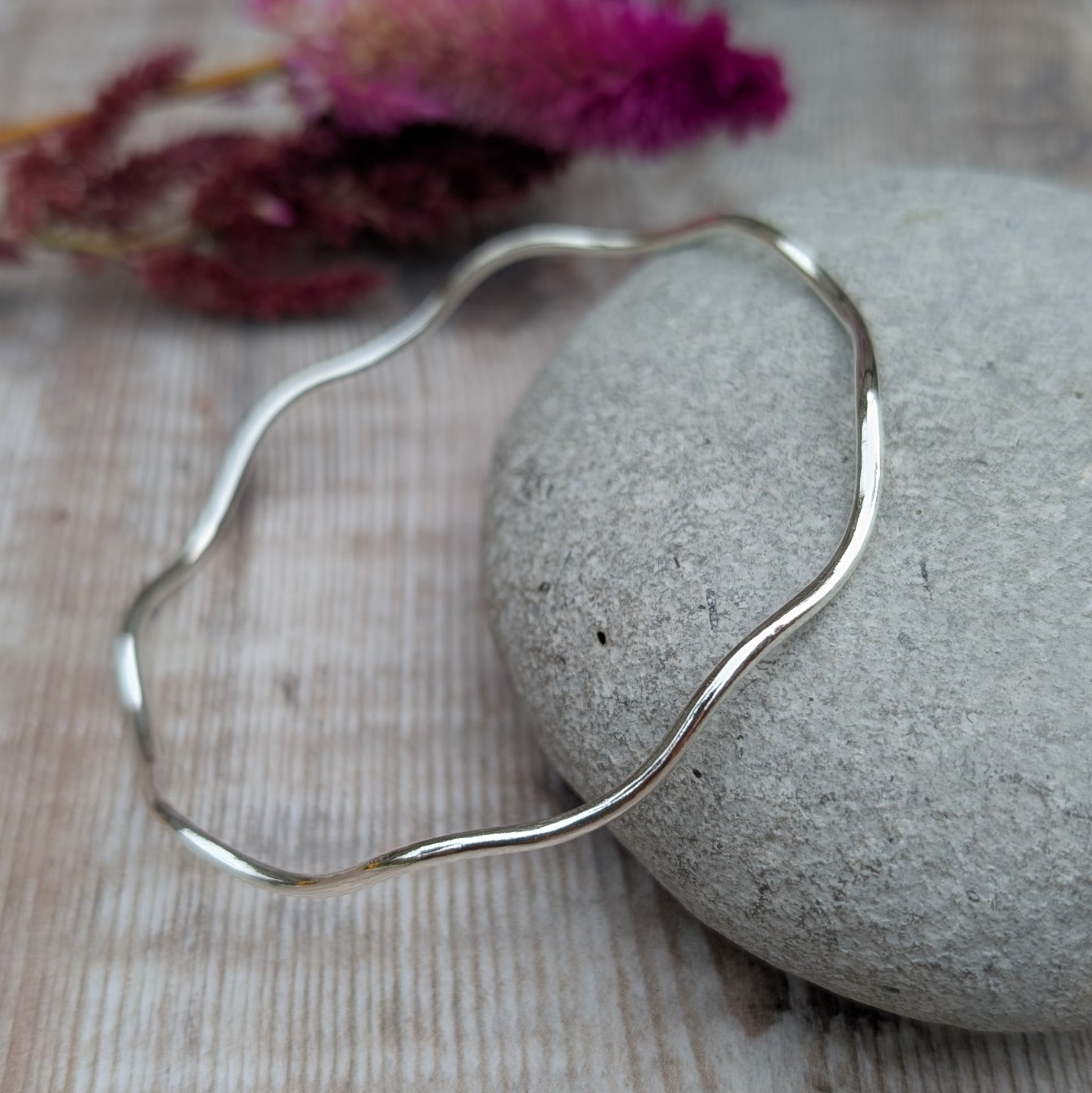 A sterling silver wave bangle resting on a smooth stone, displaying its elegant undulating design and polished surface.