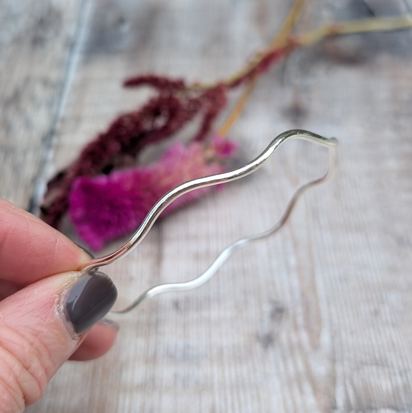 A handmade sterling silver wavy bangle held between fingers, showcasing its organic curves and polished finish against a rustic wooden background with dried flowers.
