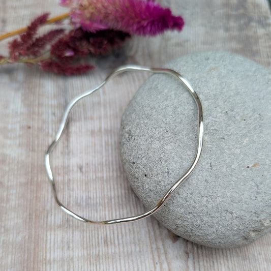 A sterling silver wave bangle resting on a smooth stone, displaying its elegant undulating design and polished surface.