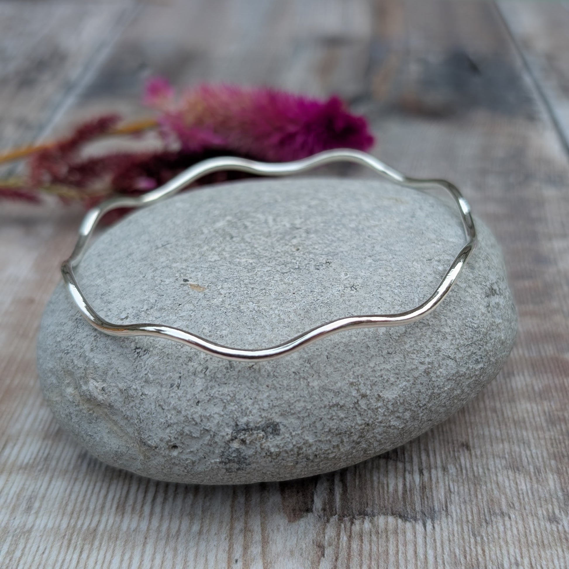 A sterling silver wave bangle resting on a smooth stone, displaying its elegant undulating design and polished surface.